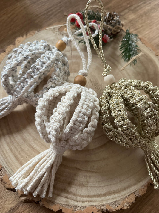 Three macrame baubles made with silver, natural and gold yarns, laying on a wooden surface next to each other. 