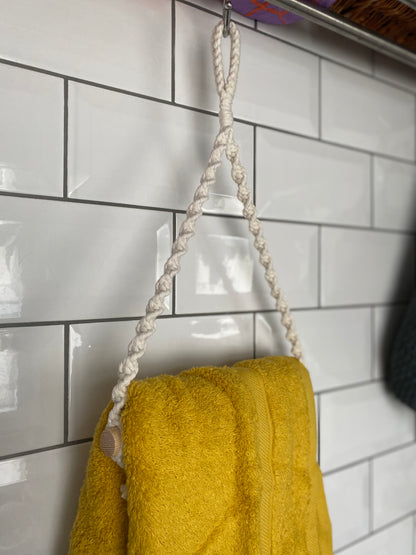 A yellow towel made from sustainable recycled cotton hangs on a white tile wall using a Macra-Made-With-Love Hand towel holder looped over a silver hook. The glossy tiles shimmer, and part of an eco-friendly woven basket is visible on a shelf above.