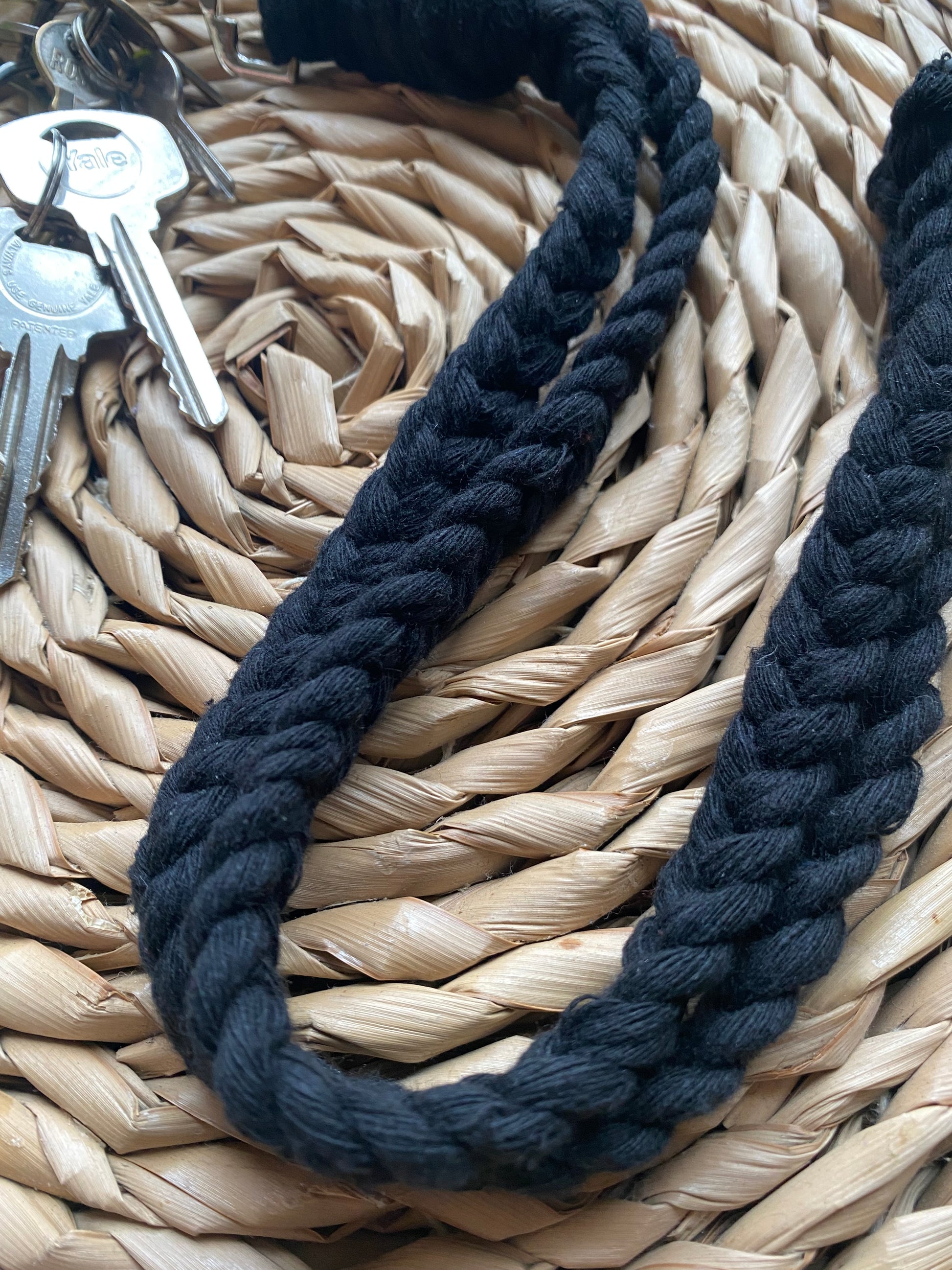 A close-up image of a set of keys with an Eco friendly lanyard badge holder - 34” by Macra-Made-With-Love lying on a woven straw surface. The keys have a metal keyring attached to the lanyard. The braided texture and pattern of the handmade lanyard are clearly visible.