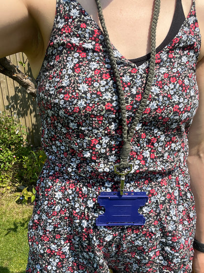 A person wearing a black floral-patterned dress with red, white, and pink flowers is standing outdoors. They have a Macra-Made-With-Love Eco friendly lanyard badge holder - 34” around their neck holding a blue ID badge holder. The background shows a wooden fence and some greenery.