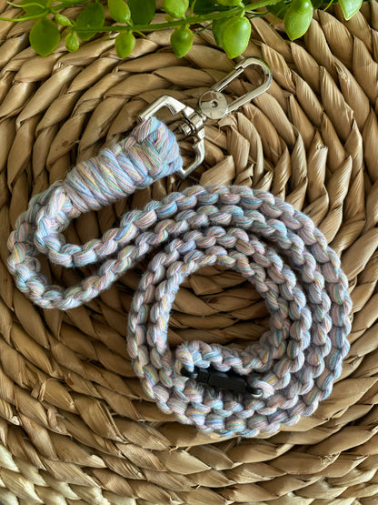 A pastel-colored, braided dog leash with a metal clasp lies coiled on a woven straw mat. In the background, there is some greenery with small, round leaves. This handmade lanyard-like design adds an eco-friendly charm to your pet's accessories. The Macra-Made-With-Love Eco friendly lanyard badge holder - 34” enhances this natural aesthetic beautifully.