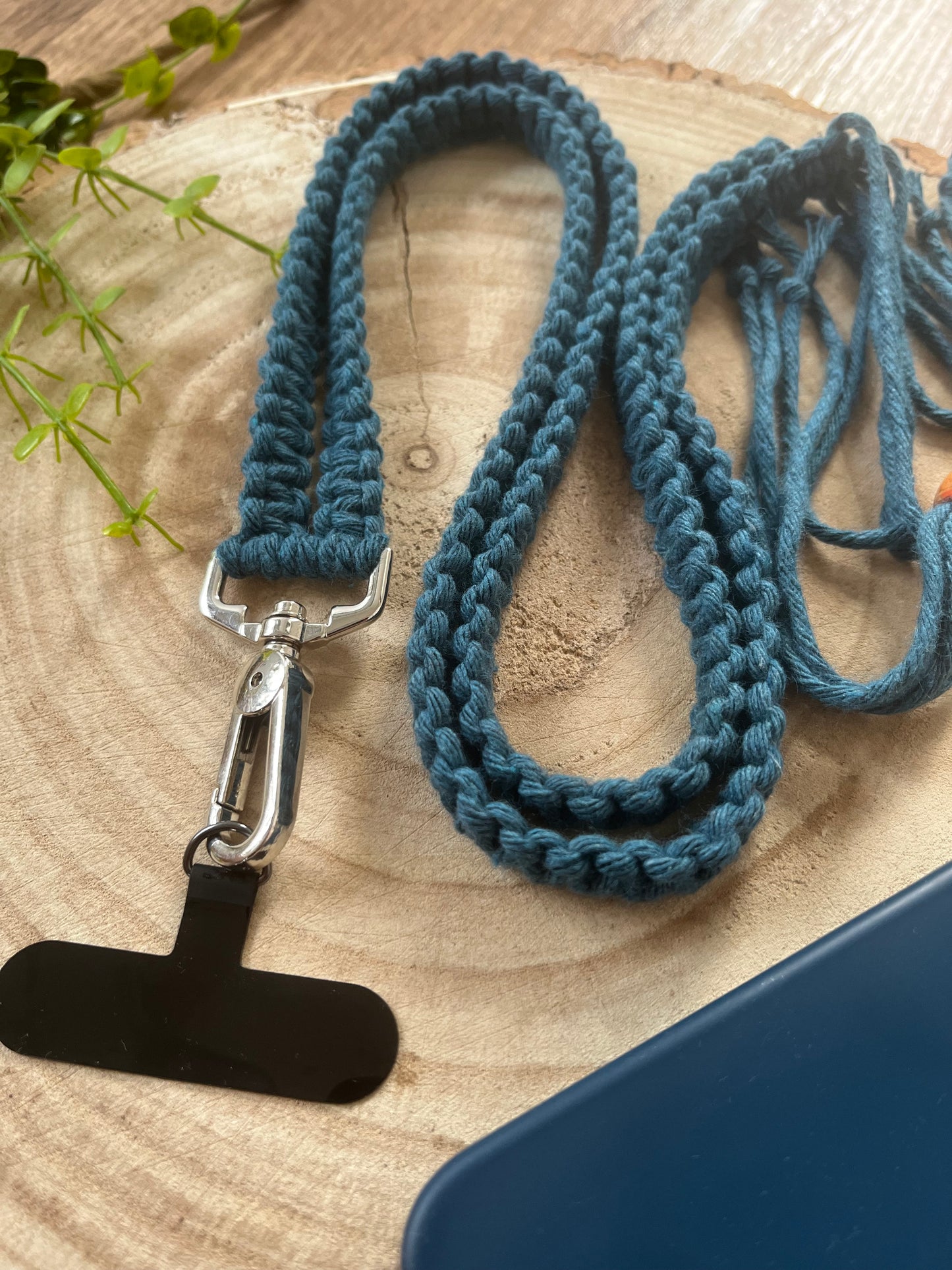 A Recycled Cotton Adjustable Crossbody Phone Strap with Universal Tether from Macra-Made-With-Love, featuring a braided blue design with a metal clasp and black attachment, lies coiled on a circular wooden surface. Adjacent objects, such as a small green plant and part of an eco-friendly blue item, are partially visible in the frame.