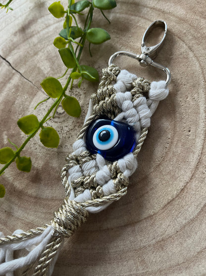 An Evil eye keychain from Macra-Made-With-Love featuring a blue evil eye charm is placed on a wooden surface. The keychain is crafted from white and beige braided cords with metallic accents. A few green leaves lie beside it, adding a natural touch to the composition.