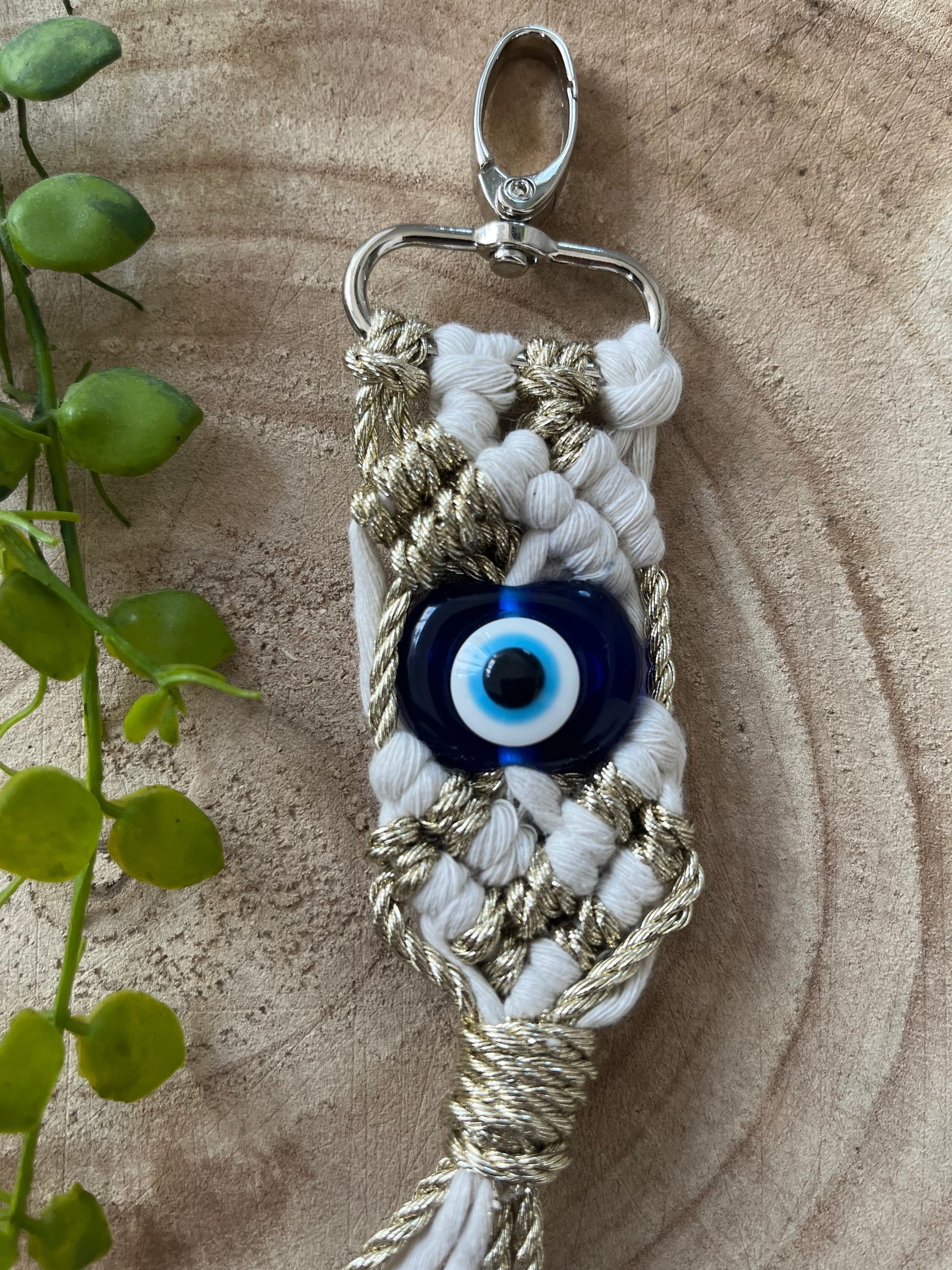 A handmade keychain with white and gold threads is pictured on a wooden surface. It features a blue evil eye bead in the center, offering protection. To the left, a small green plant adds a touch of natural contrast to the scene. The Evil Eye Keychain by Macra-Made-With-Love combines elegance and cultural significance beautifully.