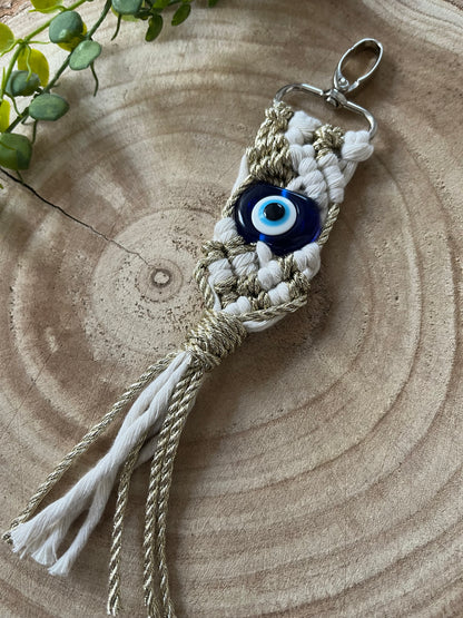 A decorative, handmade Evil Eye keychain crafted with beige and white macramé knots, featuring a blue and white bead resembling an evil eye. The Macra-Made-With-Love Evil Eye Keychain has a silver clasp at the top and tassels at the bottom, placed on a circular wooden surface with green leaves in the background.