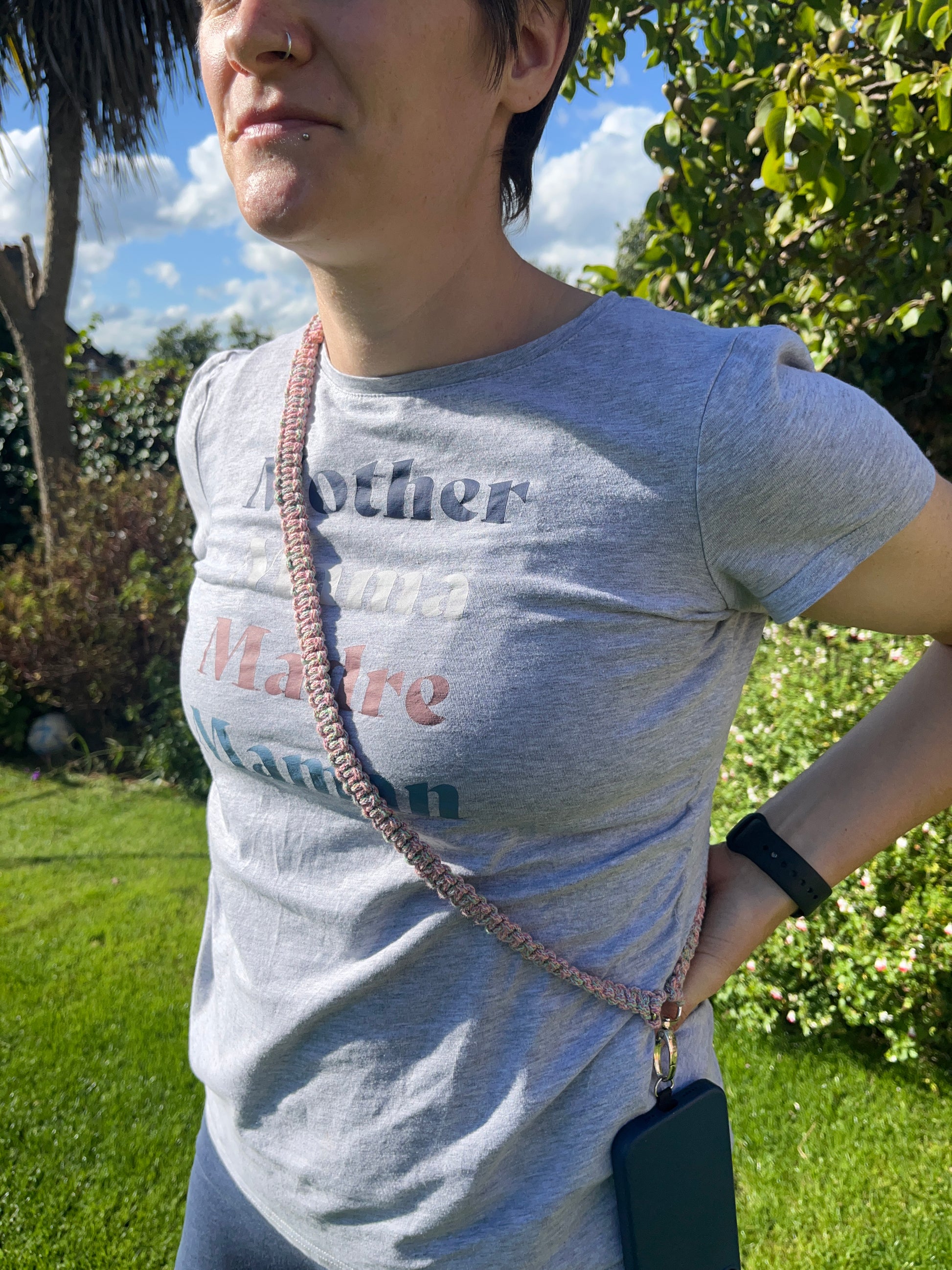 A person stands outdoors on a sunny day, wearing a gray T-shirt with the words "Mother, Mama, Madre, Maman" in colorful text. They have a Recycled Cotton Adjustable Crossbody Phone Strap with Universal Tether from Macra-Made-With-Love made from braided cord and a smartwatch on their wrist. Trees and a blue sky can be seen in the background.