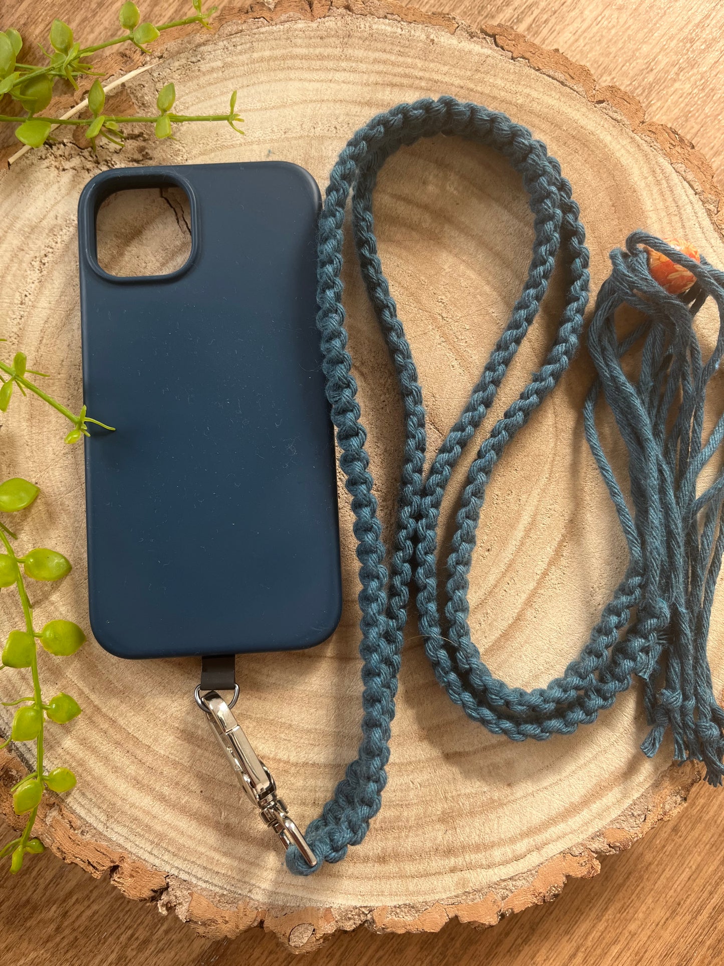 A Macra-Made-With-Love Recycled Cotton Adjustable Crossbody Phone Strap with Universal Tether, featuring a dark blue phone case and a teal braided lanyard, is placed on a wooden surface. The eco-friendly accessory complements green plant leaves situated on the left side of the surface.