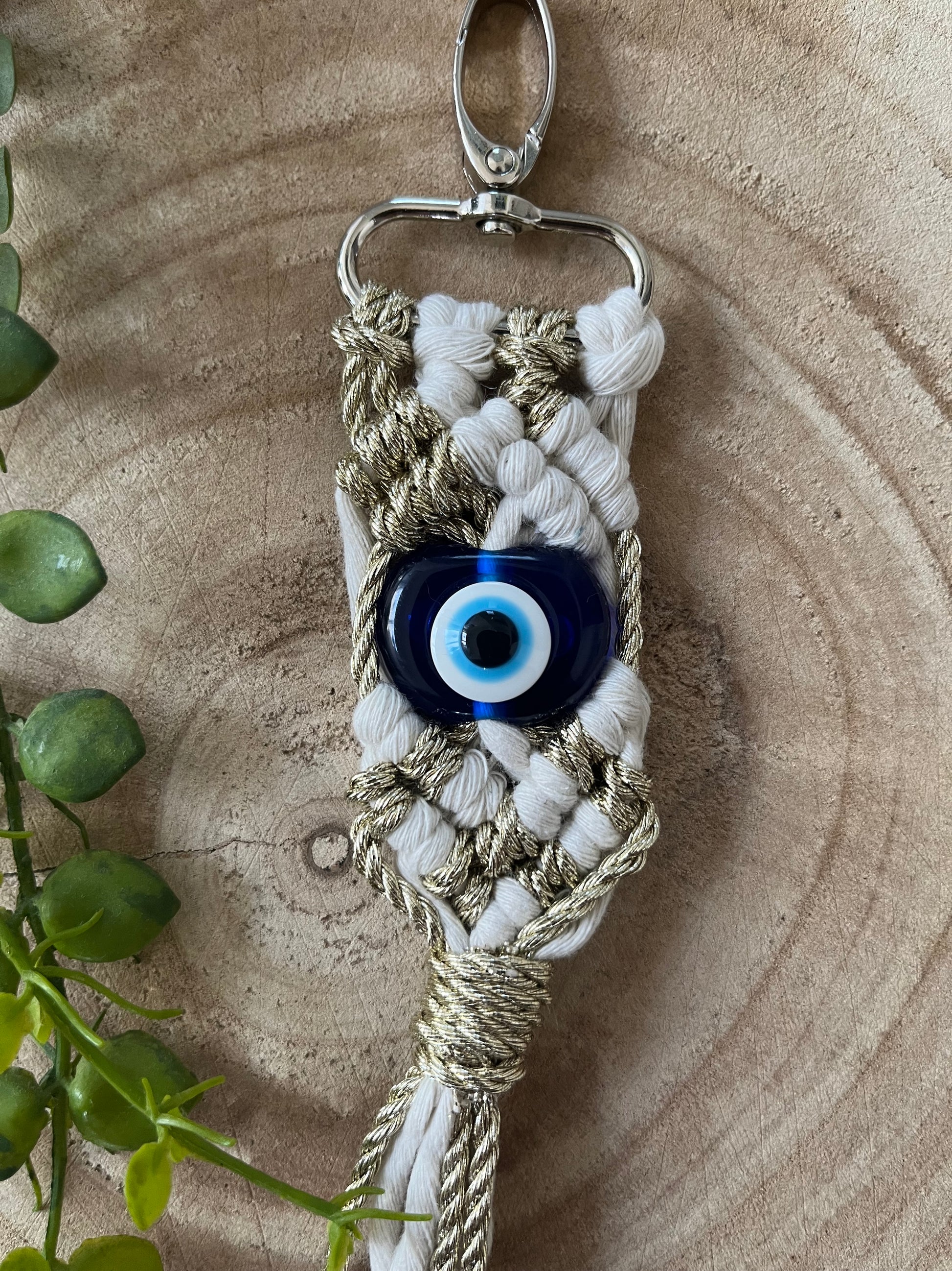 A handmade keychain made of white and gold macrame with an attached blue Evil Eye charm lies on a wooden surface. A small clipping of green plant leaves is visible in the lower left corner, enhancing the beauty of this Macra-Made-With-Love Evil eye keychain.