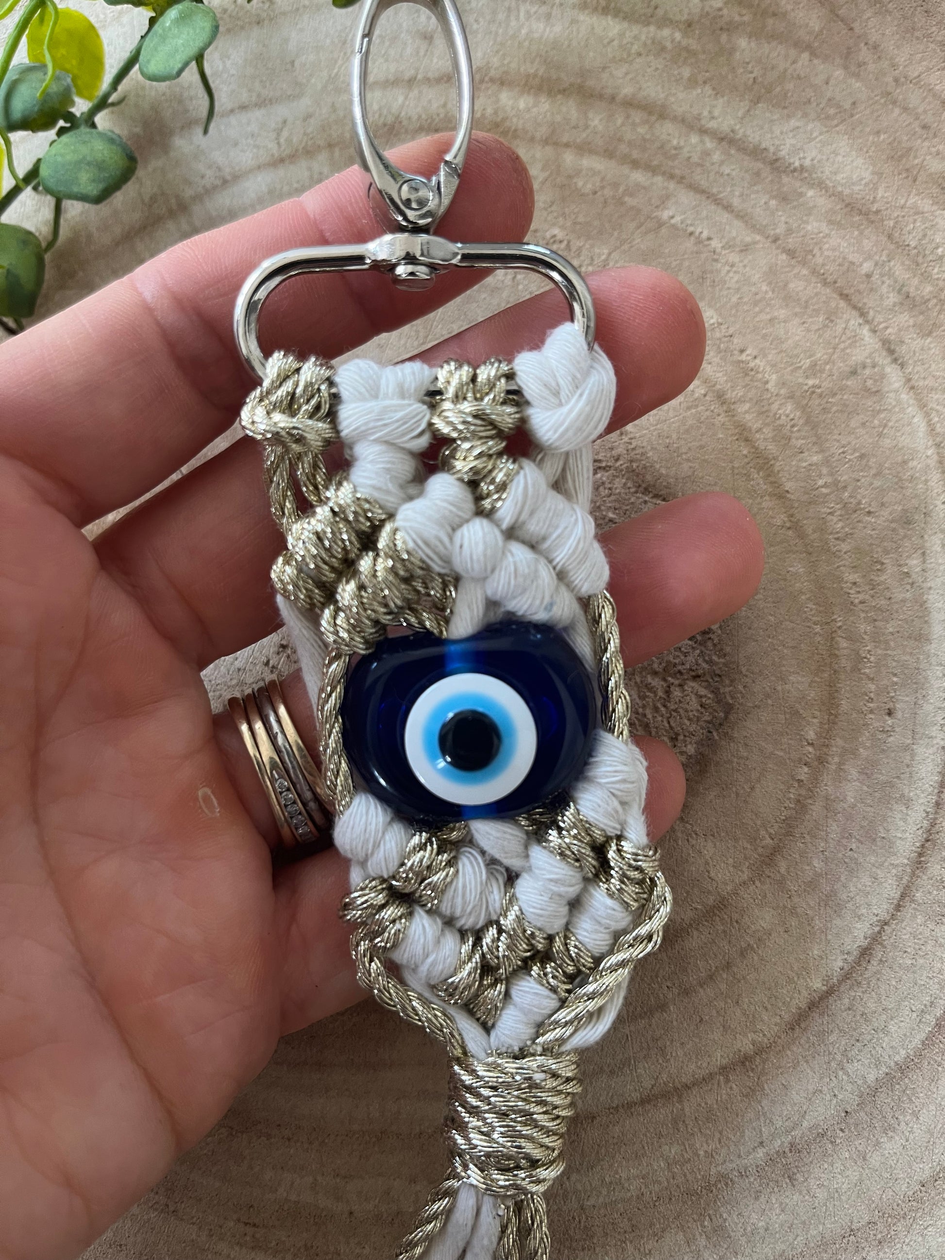 A hand holds a handmade Evil eye keychain featuring an intricate white and gold macrame design. At the center of the Macra-Made-With-Love keychain is a blue Evil Eye bead. The background is a circular wooden pattern with a green plant partially visible in the upper left.