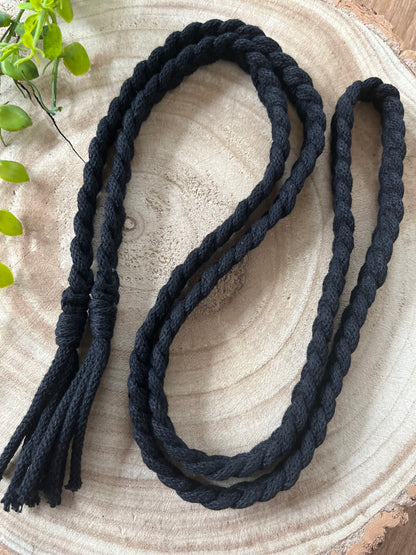 A **Braided macrame belt** by **Macra-Made-With-Love**, with tassels on one end, is coiled neatly on a wooden surface. A small green plant is partially visible in the upper left corner, adding a touch of color to the natural wooden background.