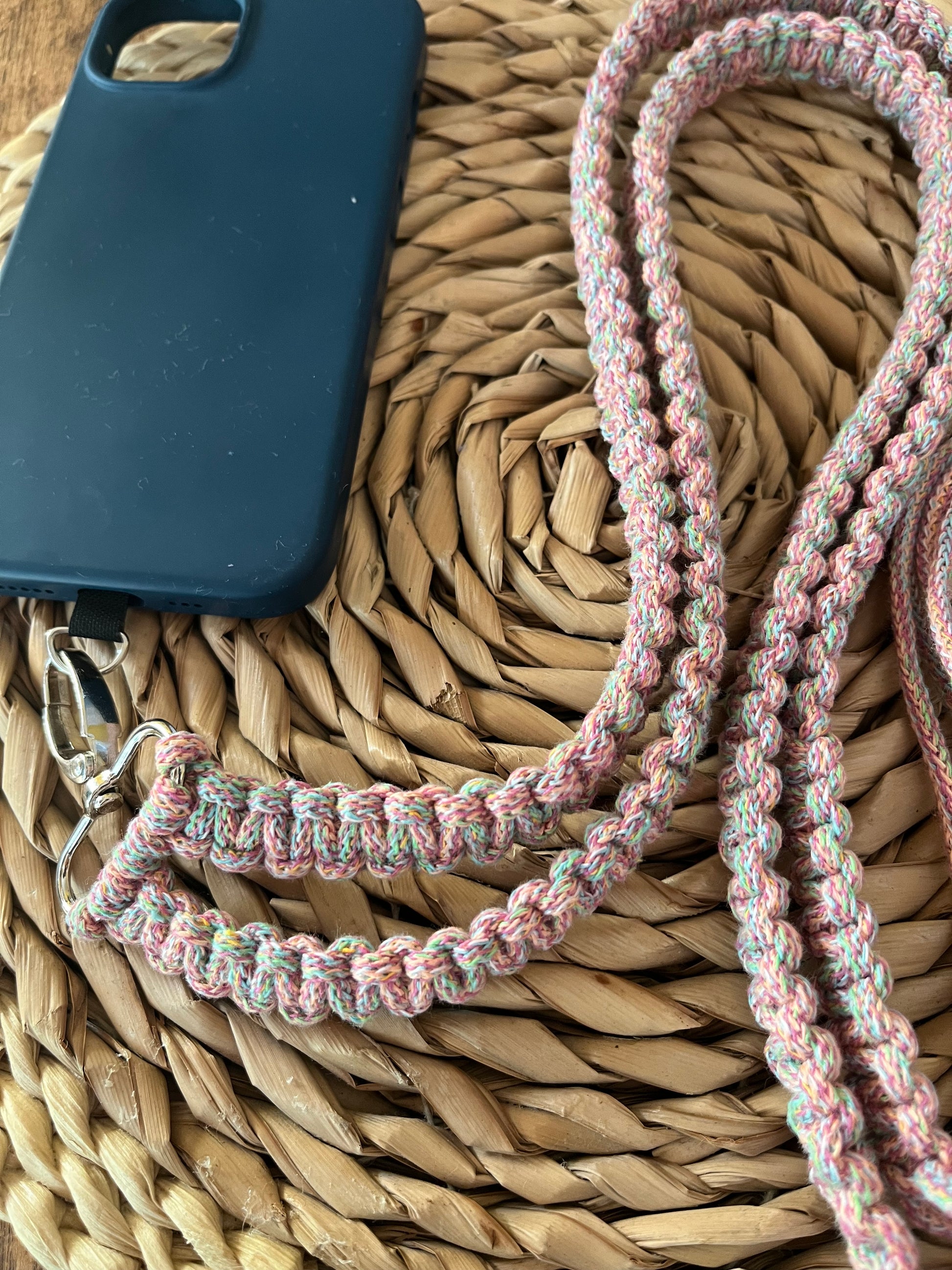 A close-up of a black phone case with an attached Recycled cotton adjustable crossbody phone strap with universal tether by Macra-Made-With-Love, lying on a woven, circular straw mat. The braided strap includes shades of pink, white, and green, and has a metal clip for attachment.