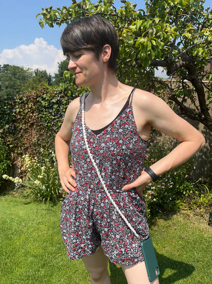 A person in a short floral romper stands in a sunny garden with their hands on their hips. They have short, dark hair and wear a smartwatch on their left wrist. A small green Macra-Made-With-Love Crossbody phone holder (double clip) made from recycled cotton cord, hanging from a white strap, is draped across their body.