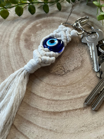 A close-up of a handmade Evil Eye keychain from Macra-Made-With-Love placed on a wooden surface. The keychain features a knotted, white macrame design with a prominent blue Evil Eye bead in the center. Several metal keys are attached to the keyring, and green plant leaves are visible in the background.