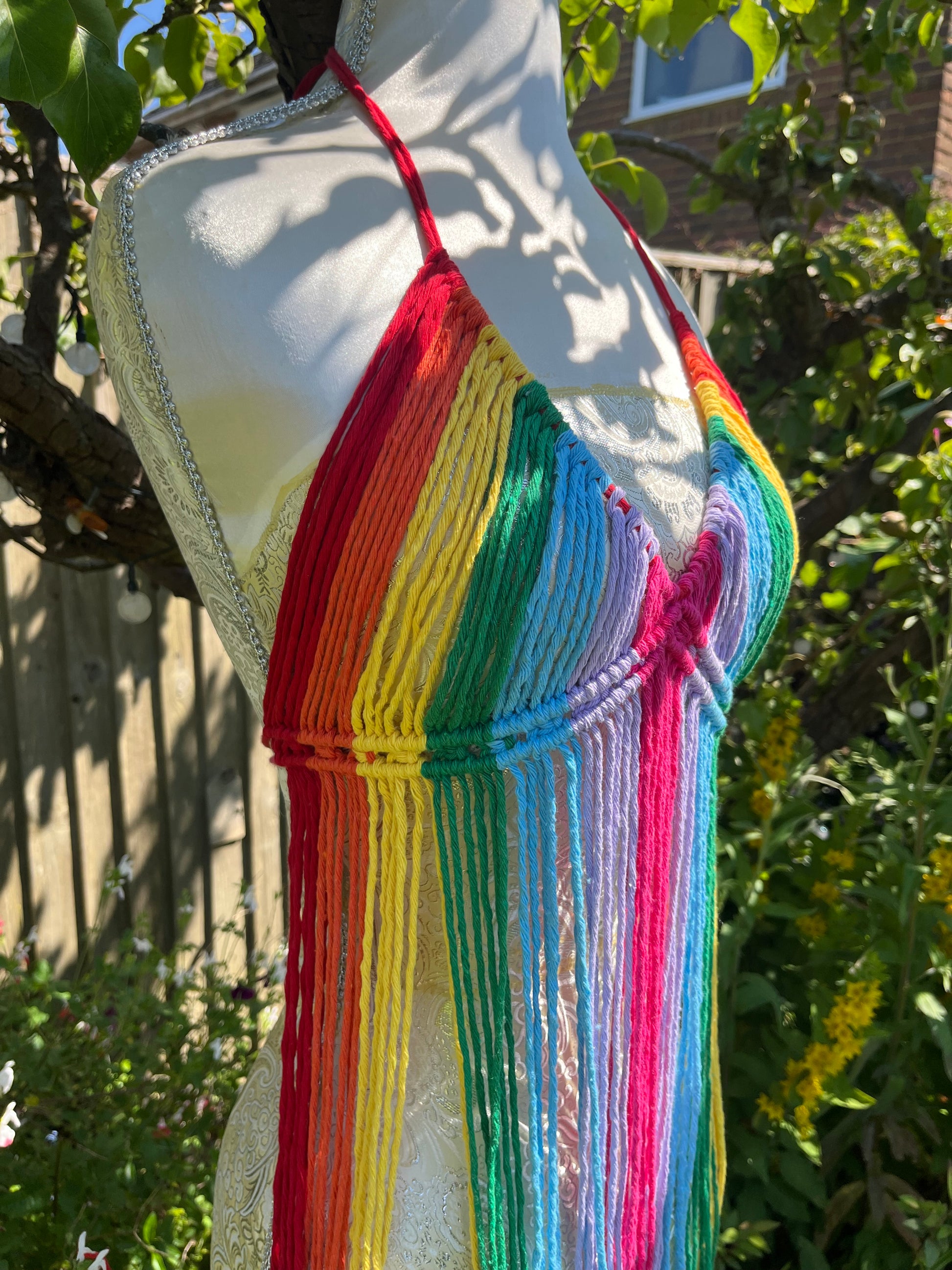 A mannequin displays a multicolored, crochet halter top with long, fringe-like strings hanging down. This Rainbow coverup top by Macra-Made-With-Love features vibrant rainbow colors arranged in vertical stripes. In the background, there are greenery, flowers, and a wooden fence. Perfect for a festival outfit!