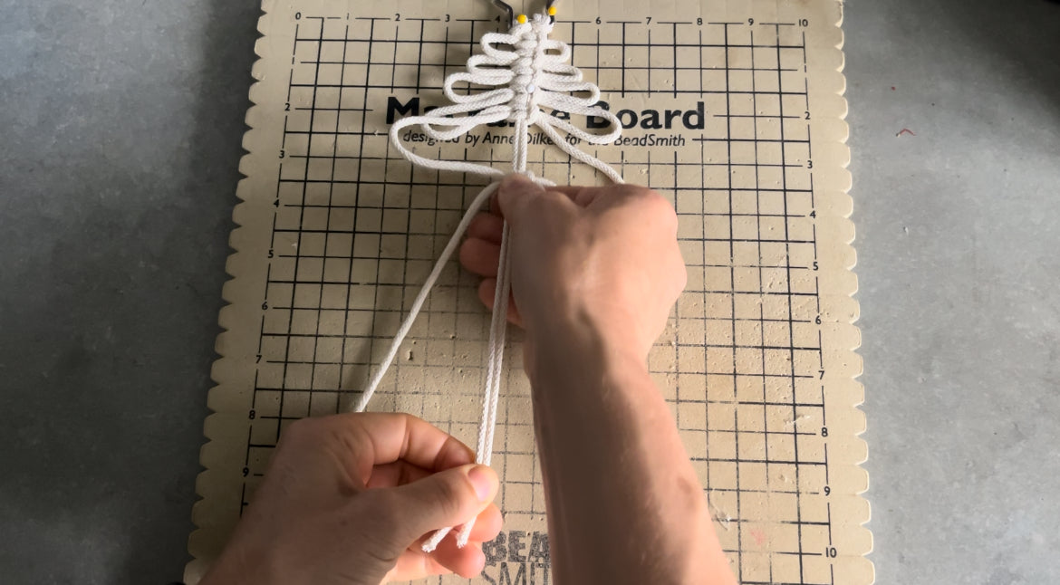 Two hands are shown crafting a Macrame Christmas Tree using white cord on a grid-patterned macramé board. The DIY tree shape is partially done and held in place with pins at the top. The background is a plain, light surface, making it perfect for enthusiasts of Macra-Made-With-Love's "Macrame Christmas Tree tutorial - PDF pattern + video" Christmas craft kit for adults.