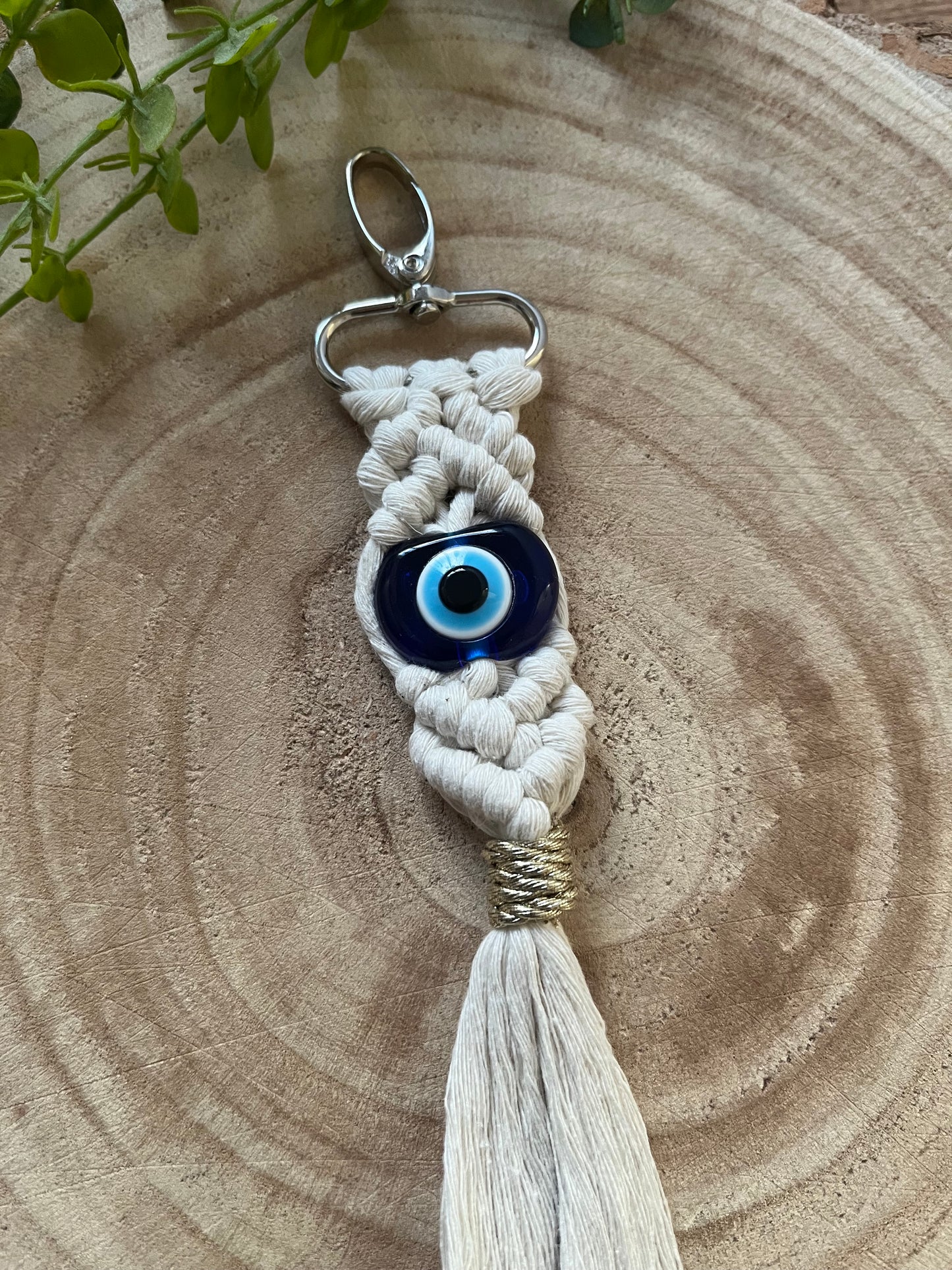 The Macra-Made-With-Love Evil eye keychain shaped like an owl features a prominent blue and white evil eye bead at its center. A long tassel hangs from the bottom. The eco-friendly keychain is placed on a round wooden surface with green foliage partially visible in the background.