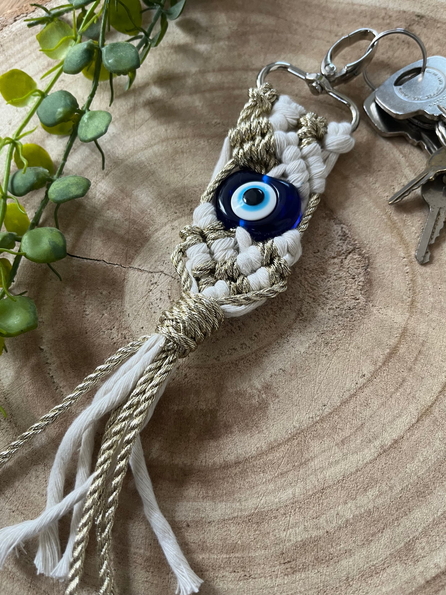 A handcrafted Evil Eye keychain with beige and white threads from Macra-Made-With-Love is adorned with an evil eye bead in the center. It lies on a wooden surface next to a few keys and a green leafy plant. This protection keychain features intricate knot work and dangling tassels at the bottom.