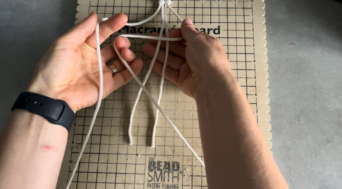 Hands are arranging white cords on a macramé board with a grid pattern, perfect for following the Macrame Christmas Tree tutorial by Macra-Made-With-Love. The board is labeled "Macramé Board" and "Beadsmith Patent Pending". The person, wearing a black smartwatch and a ring on the left hand, is working on a grey surface.