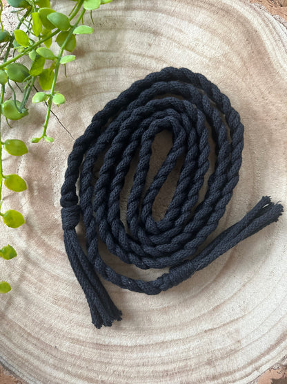 A coiled black cotton rope, reminiscent of a Braided macrame belt by Macra-Made-With-Love, rests on a round wooden surface. The thick braided texture of the rope is clearly visible. A small green plant with several leaves is partially visible in the top left corner, adding a touch of color to the composition.