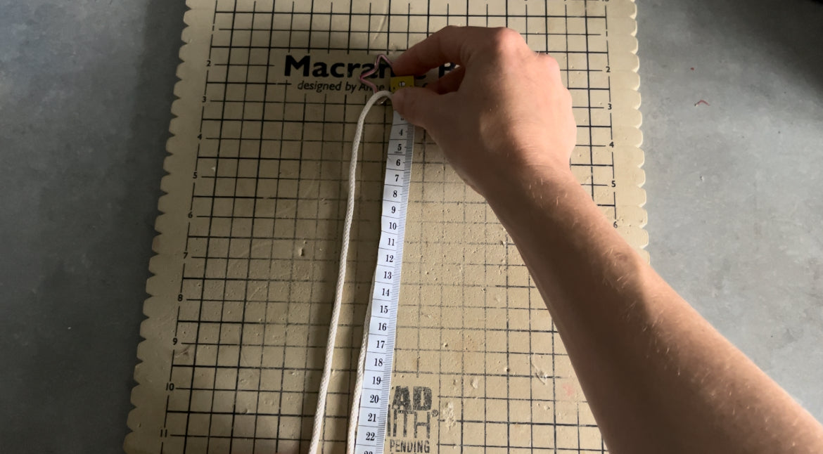 A hand is holding and measuring a piece of white rope with a flexible measuring tape against a gridded macrame board from the Macrame Christmas Tree tutorial - PDF pattern + video by Macra-Made-With-Love. The macrame board, ideal for following this tutorial, has a grid and scalloped edges. The hand appears to be above a metal surface.