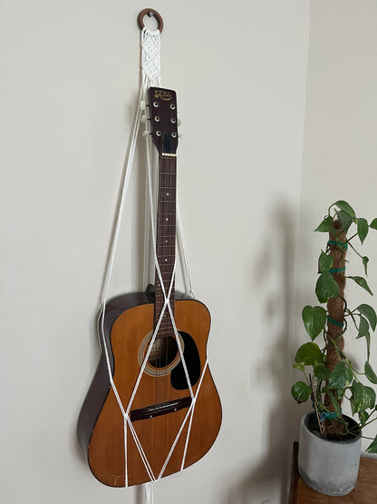 A wooden acoustic guitar is hanging on a light-colored wall using a Macra-Made-With-Love Guitar wall mount with intricate macramé knots, adding a touch of boho chic home decor. Next to it is a small potted plant with green leaves climbing a moss pole.