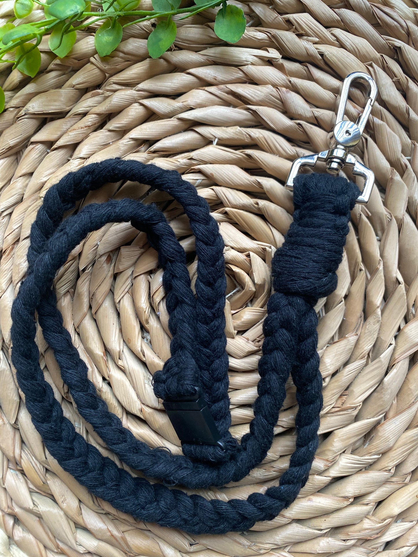 A coiled black braided rope dog leash with a metal clasp at one end is placed on a woven straw mat. A small portion of green leaves is visible in the upper-left corner of the image, complementing the natural look of this **Eco friendly lanyard badge holder - 34”** by **Macra-Made-With-Love** for your pet.