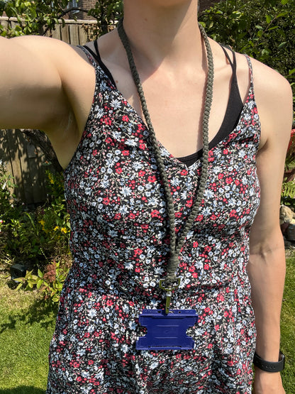 A person wearing a sleeveless floral dress with black, red, and white patterns stands outside in a garden. They have an Eco friendly lanyard badge holder - 34” from Macra-Made-With-Love around their neck with a blue ID badge holder attached. The person's face is not visible.