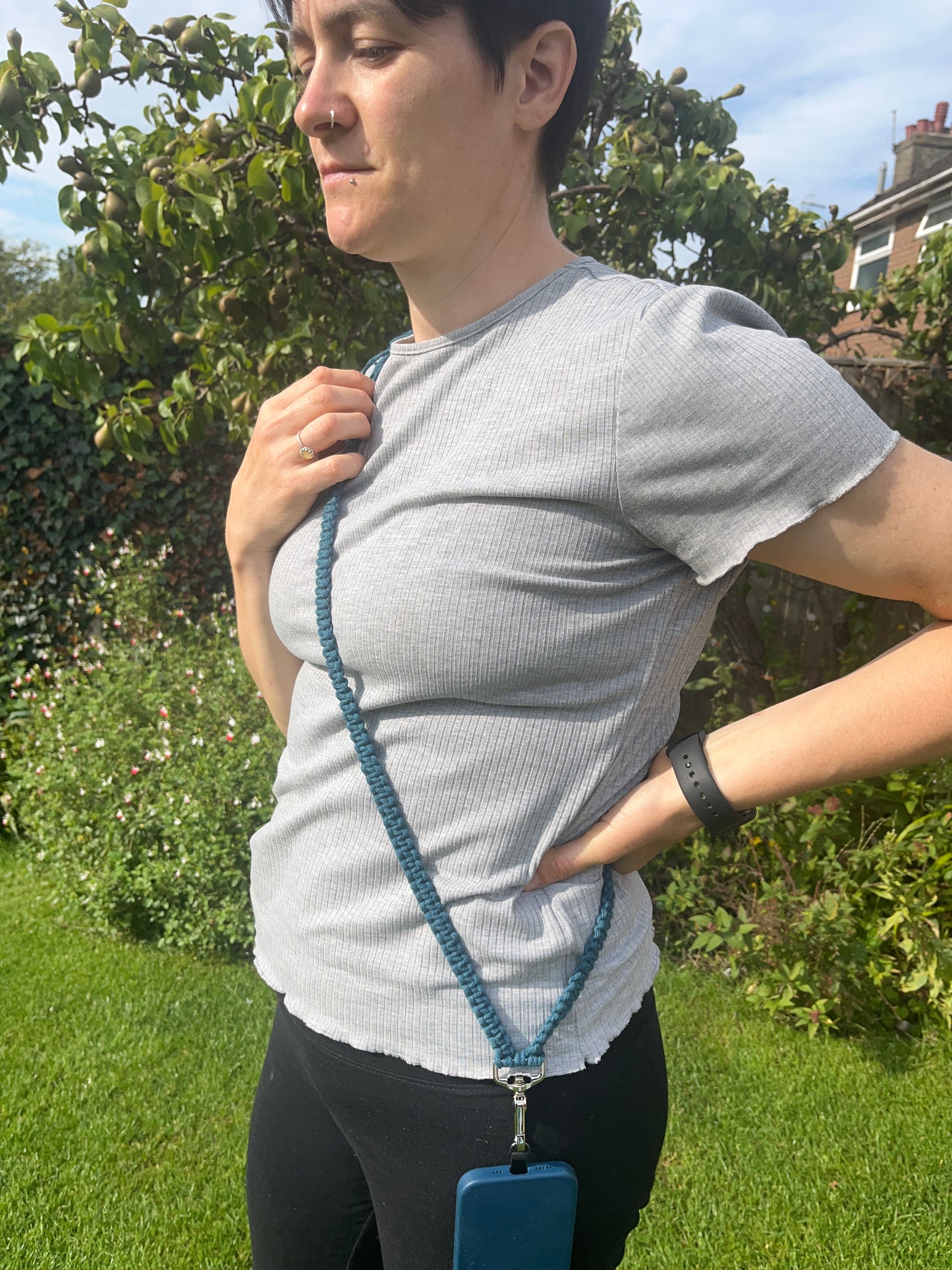 A person with short hair stands outdoors in a garden, wearing a grey ribbed t-shirt and black pants. They have the Recycled cotton adjustable crossbody phone strap with universal tether by Macra-Made-With-Love draped across their torso, attached to a phone at the end. They are looking to the side, with one hand resting on their hip.