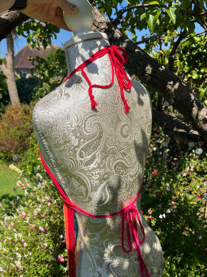 A decorative mannequin with a white paisley pattern stands outdoors, wrapped in red string. The mannequin is placed against a backdrop of lush green plants and trees, with a clear blue sky visible in the background. A hand is adjusting its neck area, as if perfecting the Rainbow coverup top by Macra-Made-With-Love.