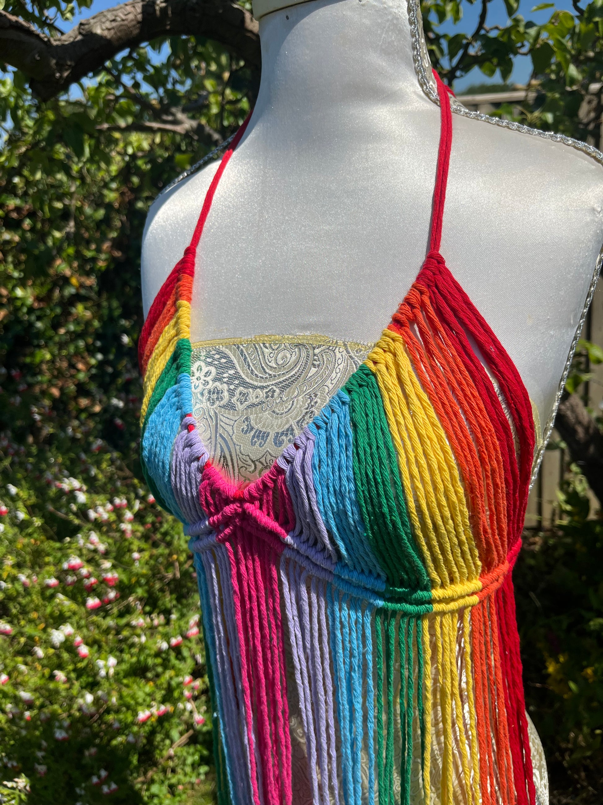 A mannequin wearing a colorful, hand-woven "Rainbow coverup top" by Macra-Made-With-Love, tied at the neck with red strings. The background shows a lush green garden with various plants and flowers under a bright, sunny sky. Perfect as a festival outfit.