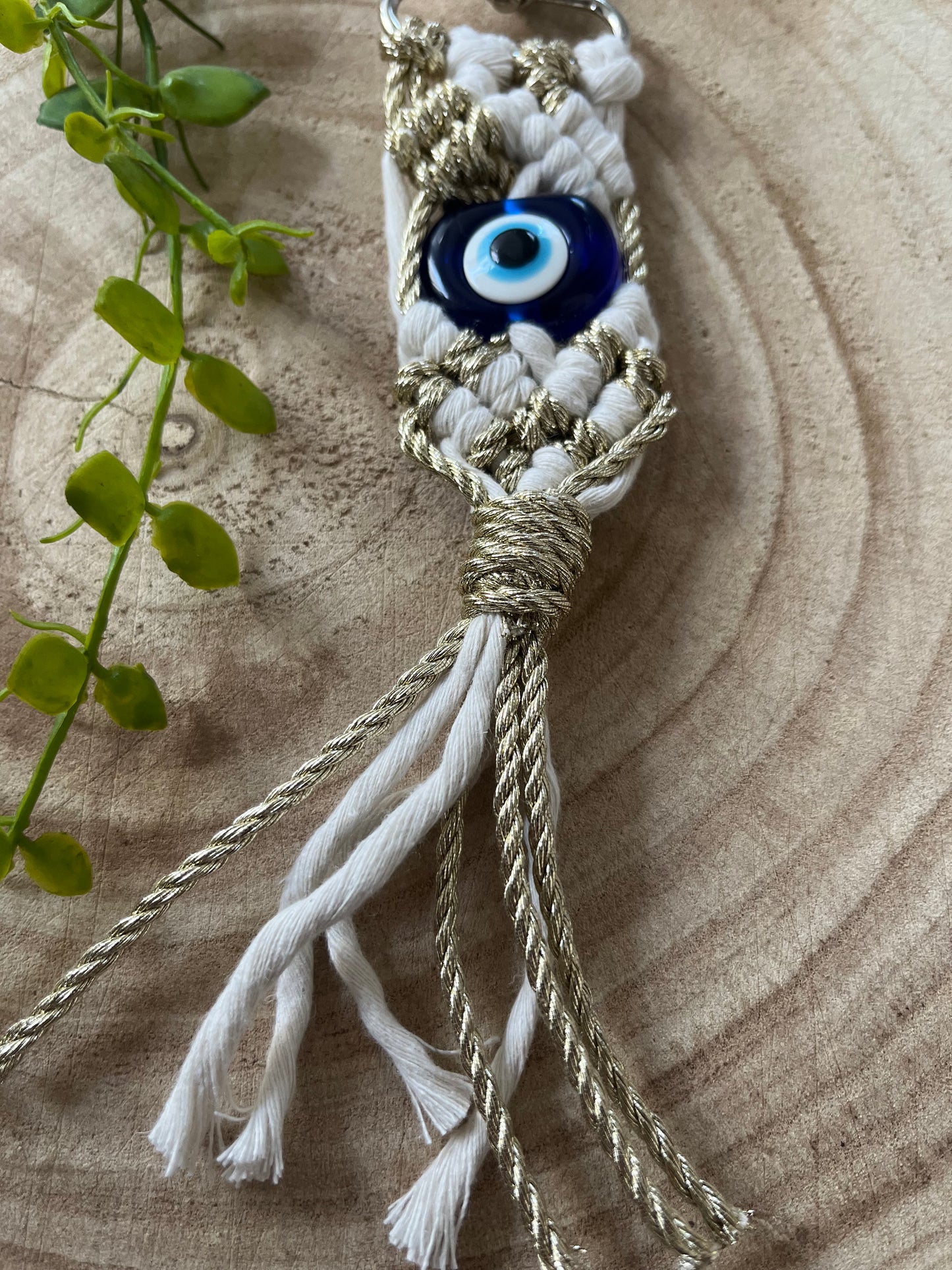 A close-up of a Macra-Made-With-Love Evil eye keychain with a blue Evil Eye bead in the center. The protection keychain features white and gold braided cords, with a few loose ends hanging at the bottom. A green plant is partially visible on the left side, placed on a wooden surface.