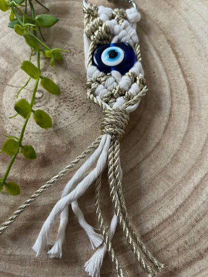 A close-up of a Macra-Made-With-Love Evil eye keychain with a blue Evil Eye bead in the center. The protection keychain features white and gold braided cords, with a few loose ends hanging at the bottom. A green plant is partially visible on the left side, placed on a wooden surface.