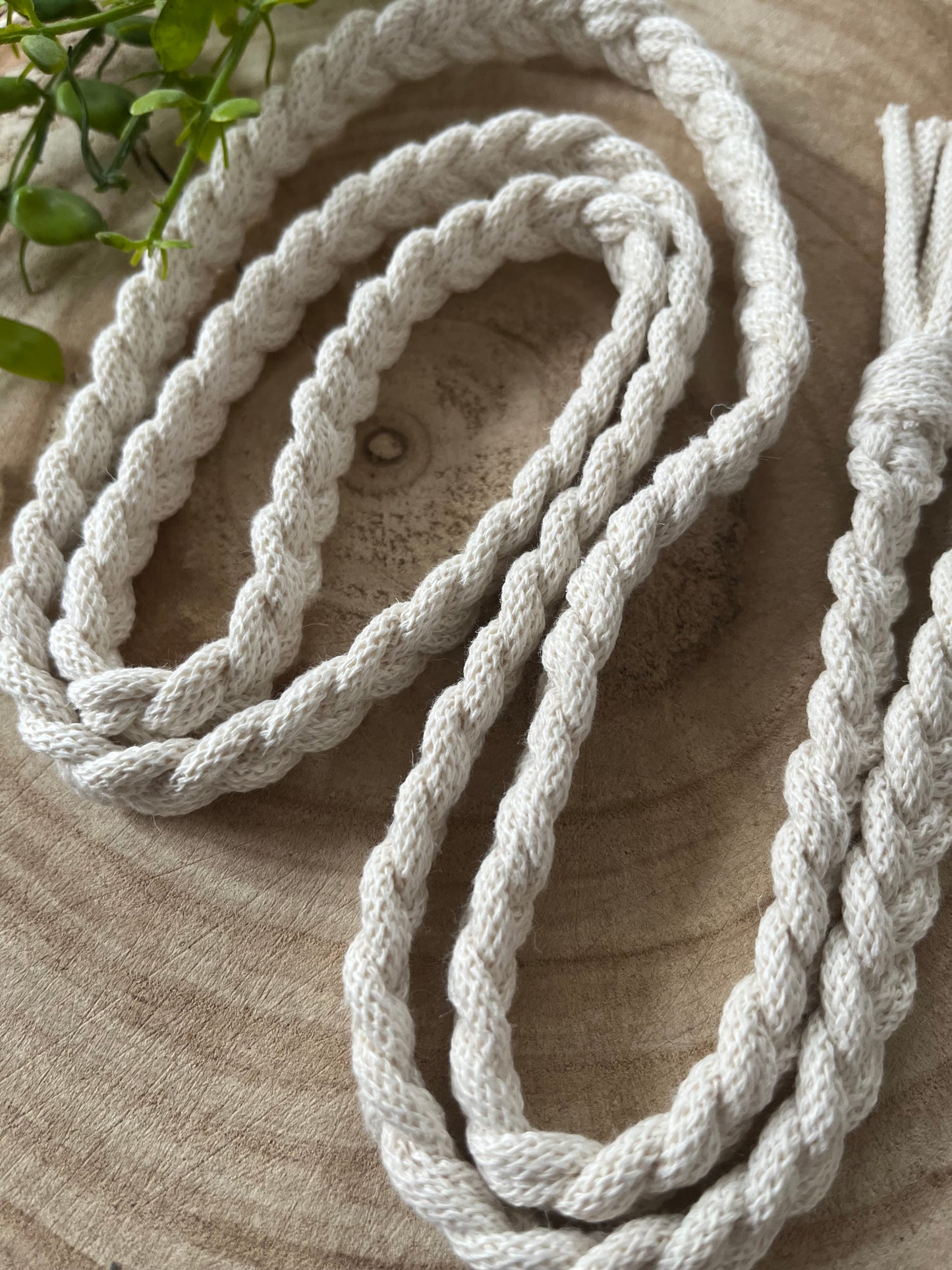 Close-up of a thick, Macra-Made-With-Love braided macrame belt coiled in a loose circular pattern on a wooden surface. In the background, green leaves partially extend into the frame from the top left corner, adding a touch of natural color to this eco-friendly composition.