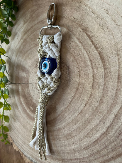 A Macra-Made-With-Love Evil eye keychain with beige and white threads braided together, featuring a blue evil eye bead in the center. This protection keychain hangs against a round wooden backdrop with a small leafy plant in the corner of the image.