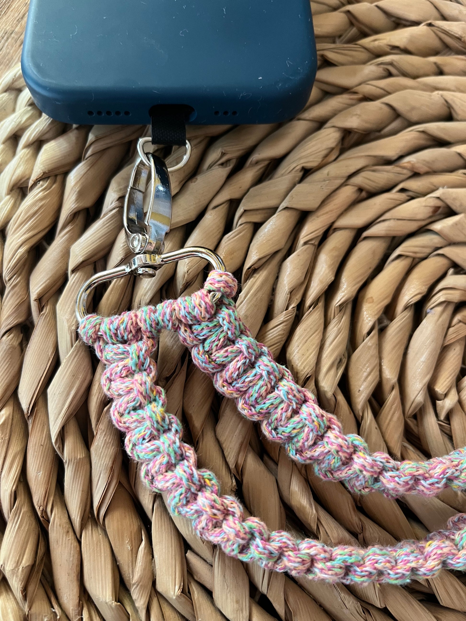 A phone with a dark blue case is attached to a keychain clip. The clip connects to the Macra-Made-With-Love Recycled Cotton Adjustable Crossbody Phone Strap with Universal Tether, an eco-friendly, handwoven, multicolored cord made from recycled cotton. The setup rests on a textured, woven surface.