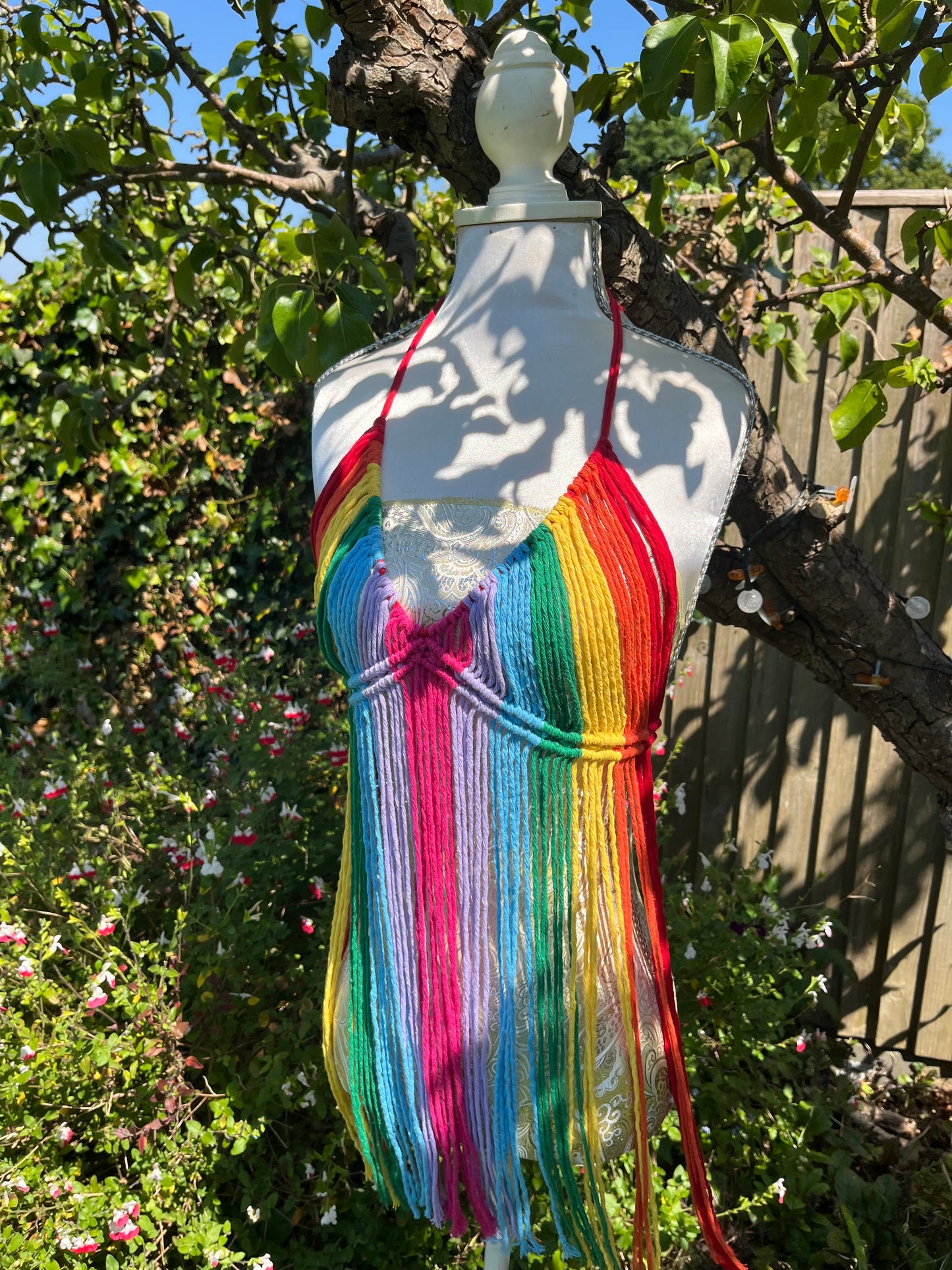 A white mannequin stands outdoors beneath a tree, showcasing the Rainbow coverup top by Macra-Made-With-Love, a vibrant, rainbow-colored crochet halter top with long, multicolored fringe cascading down the front. The festival outfit exudes eco-friendly craftsmanship, set against lush green foliage and sunlight filtering through the leaves.