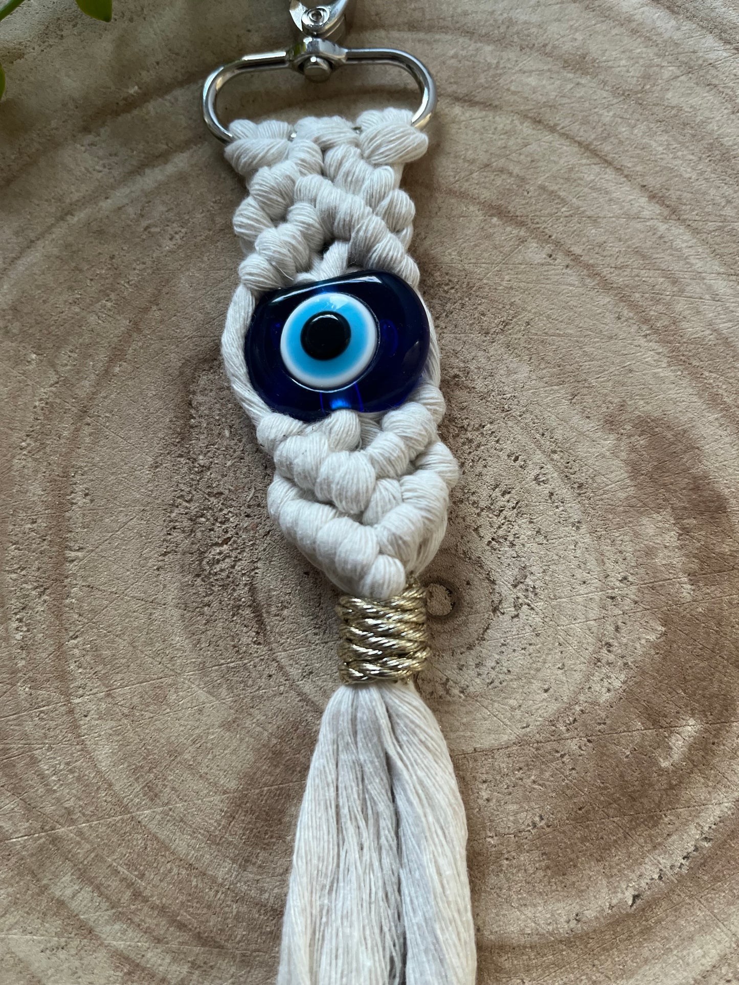 A handmade Macra-Made-With-Love Evil Eye keychain with a blue bead is displayed against a wooden background. The keychain features intricately woven white cords and ends in a tassel bound with gold thread, creating a blend of rustic and bohemian aesthetics.