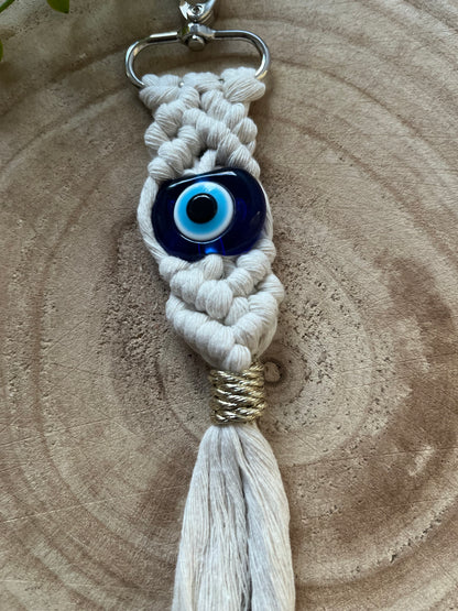 A handmade Macra-Made-With-Love Evil Eye keychain with a blue bead is displayed against a wooden background. The keychain features intricately woven white cords and ends in a tassel bound with gold thread, creating a blend of rustic and bohemian aesthetics.