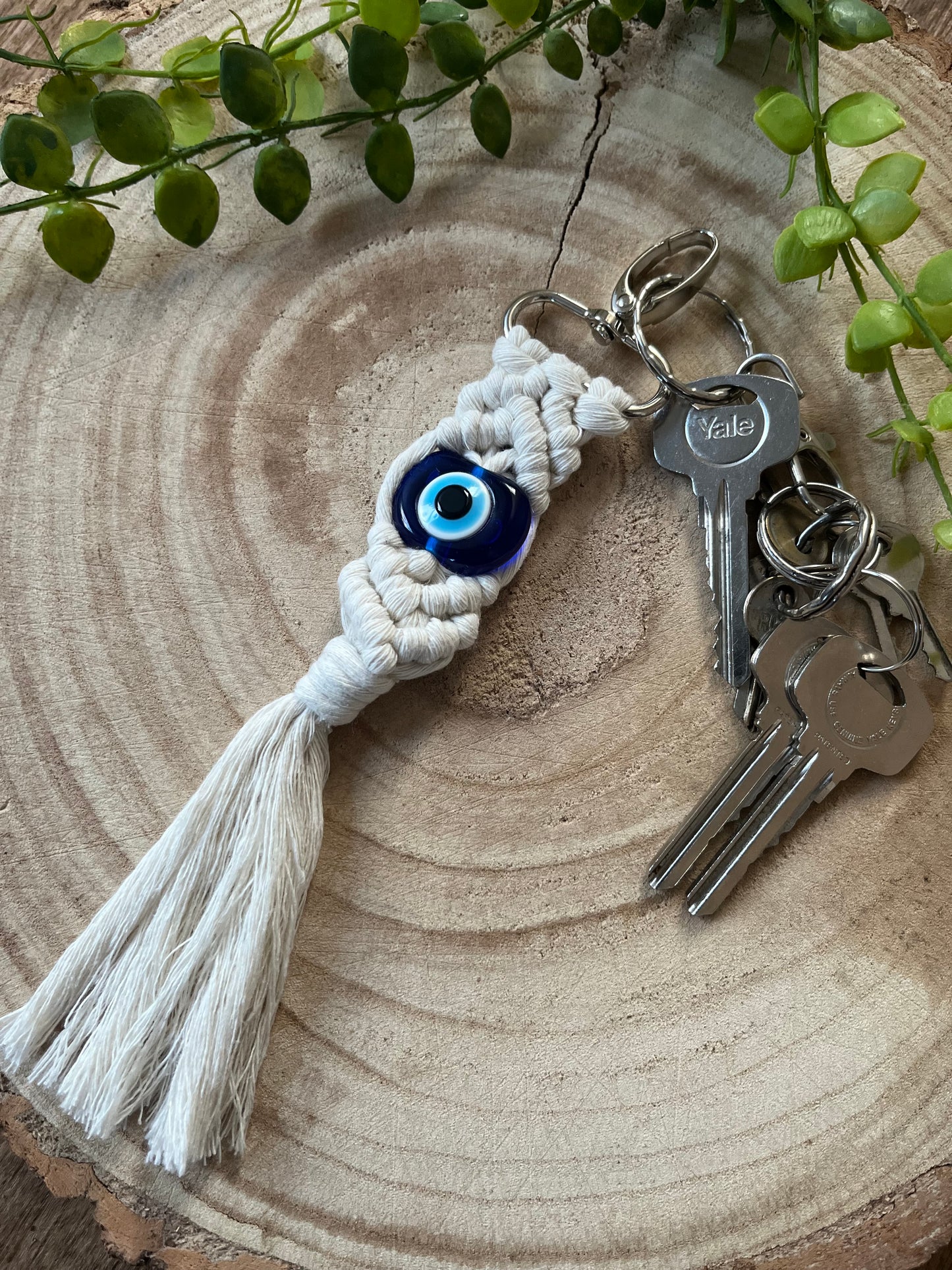 A set of keys with a Yale keychain rests on a circular wooden surface. Attached to the keys is a white macrame Evil Eye Keychain from Macra-Made-With-Love, serving as a protection talisman. Green leafy plant sprigs encircle the wooden surface, adding a natural touch to the scene.