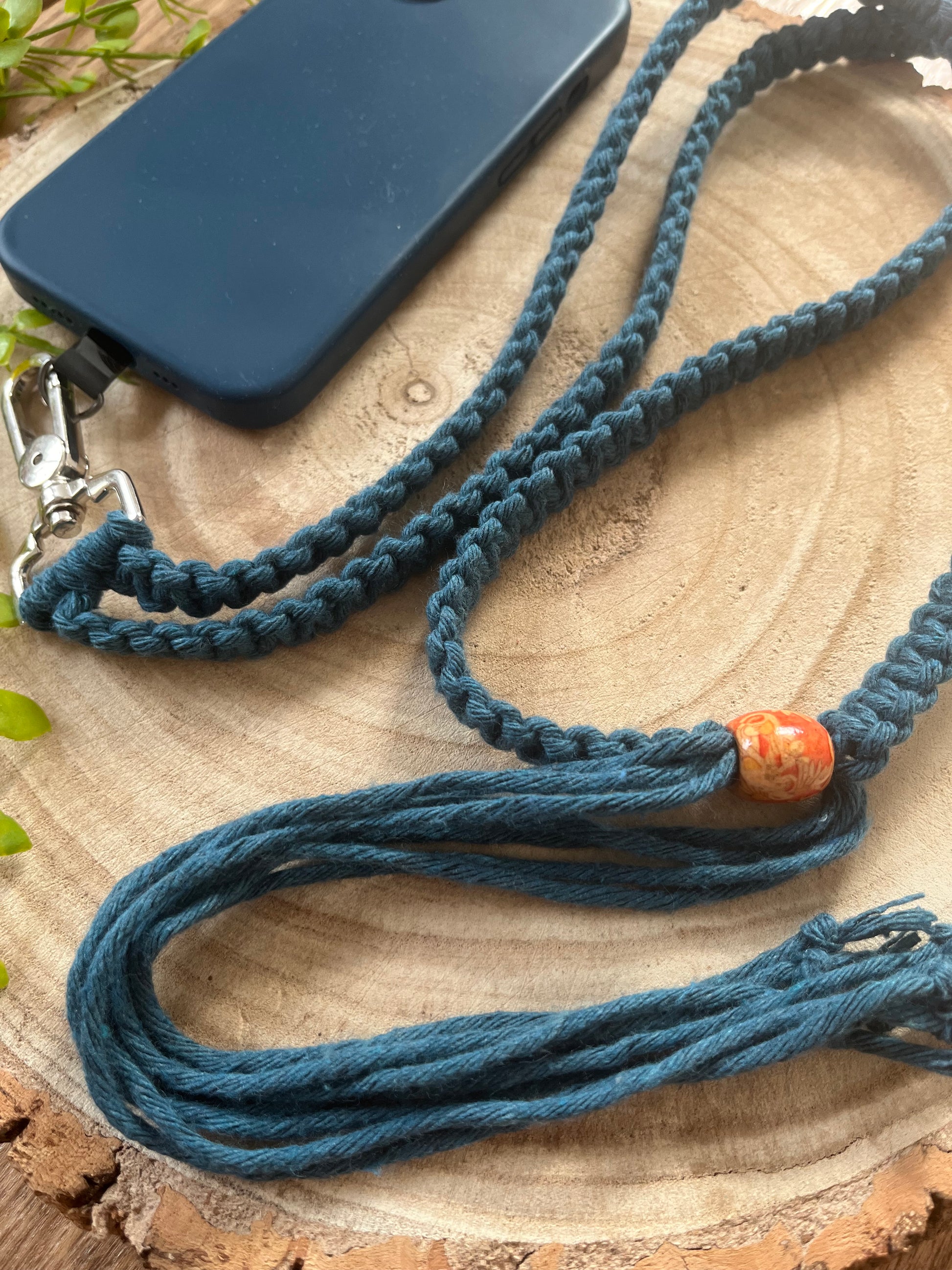 A close-up image features the navy blue braided Recycled Cotton Adjustable Crossbody Phone Strap with Universal Tether by Macra-Made-With-Love, accented with a metal clip and an amber-colored bead. This eco-friendly lanyard, crafted from recycled cotton, is attached to a dark blue phone case resting on a wooden surface. Green leaves are partially visible in the background.