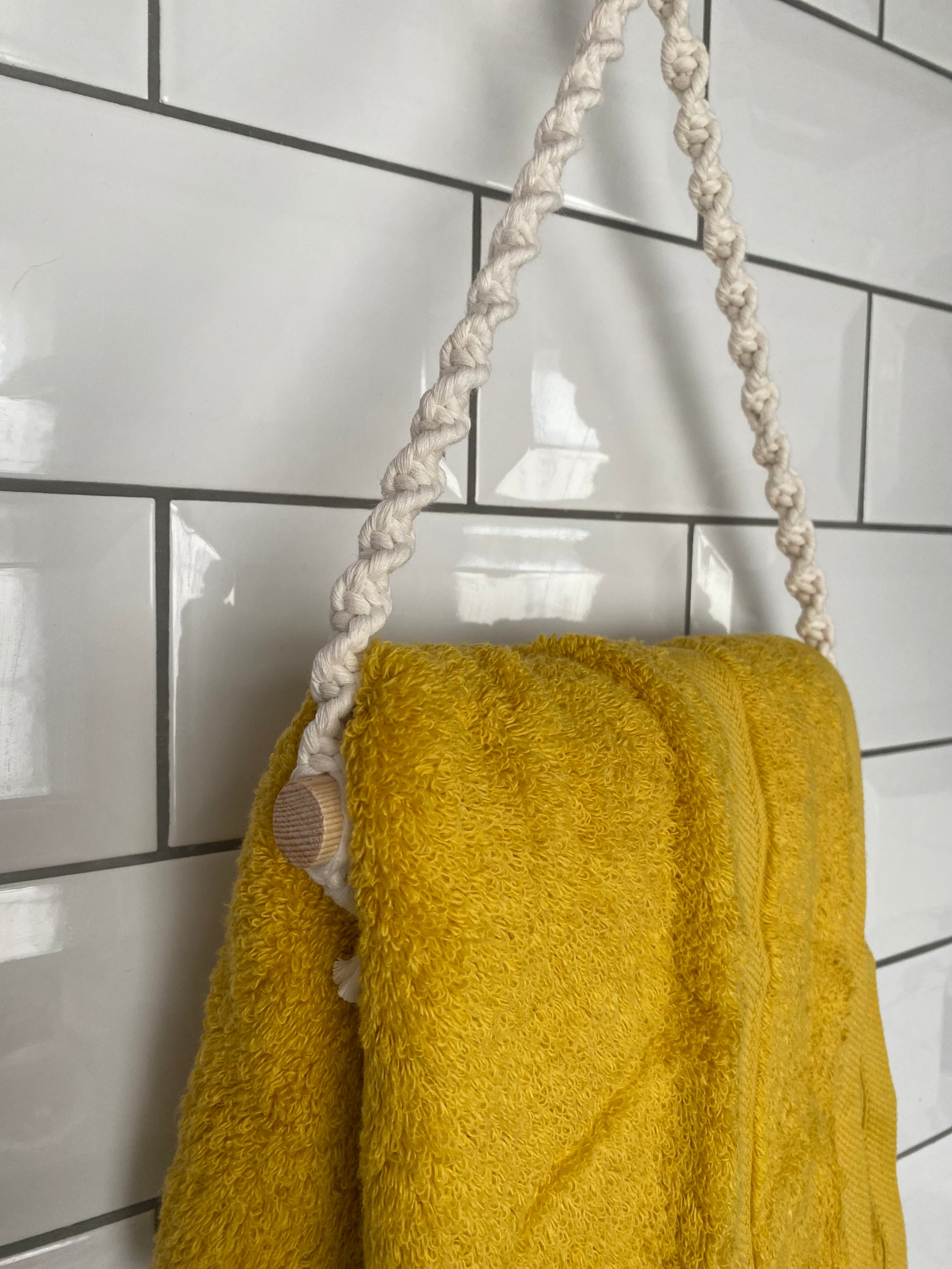 A yellow towel hangs on a **Hand towel holder by Macra-Made-With-Love**, made from sustainable recycled cotton, attached to a white tiled wall. The rope is knotted in a decorative pattern, adding an artistic touch to the practical setup. The tiles have a glossy finish and are arranged in a horizontal subway pattern.