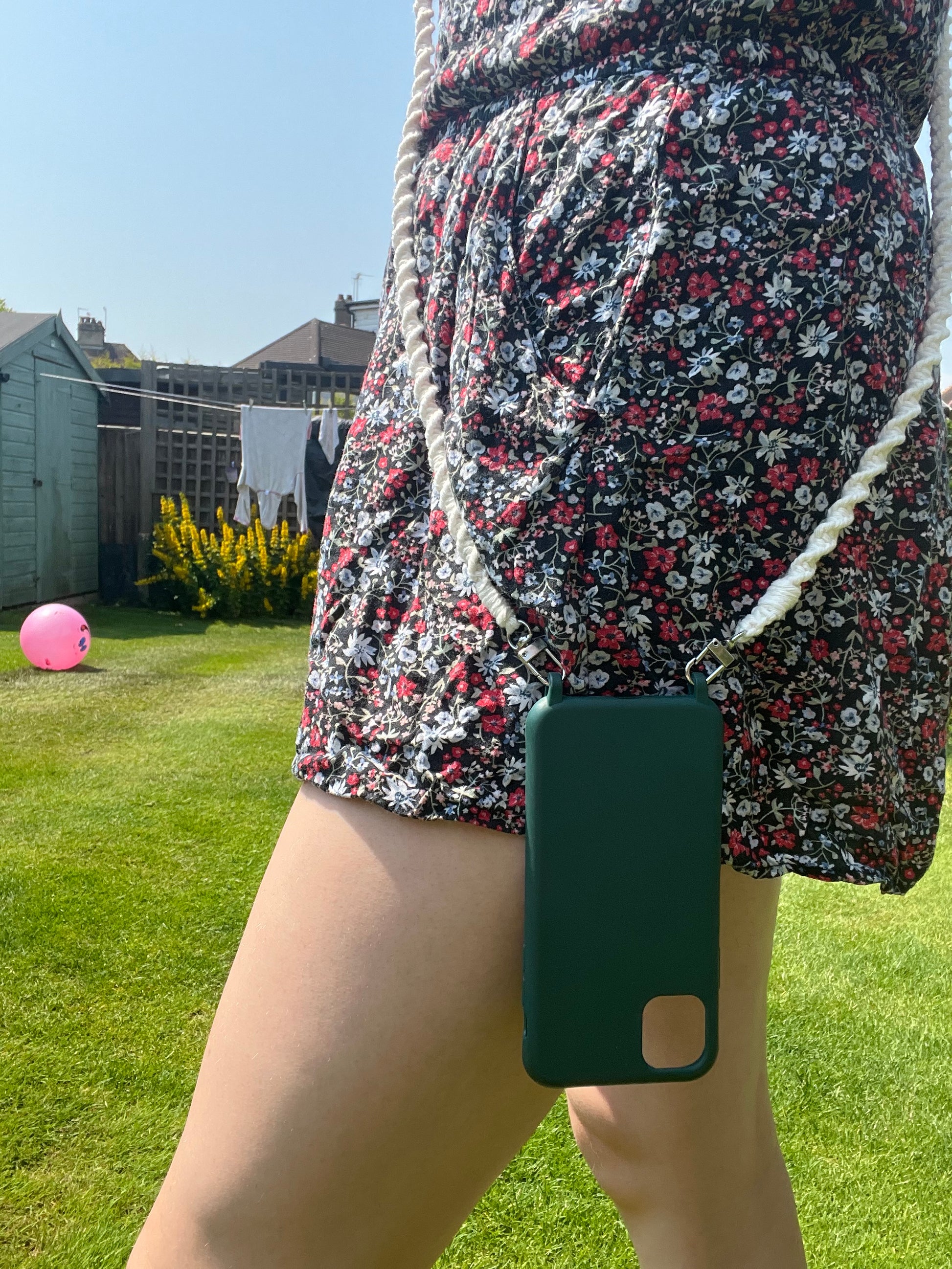 A person wearing a floral print dress stands on a grassy lawn with an eco-friendly Crossbody phone holder (double clip) by Macra-Made-With-Love featuring a black case and recycled cotton cord in white. In the background, there is a wooden shed, a fence, yellow flowers, and a pink ball. The scene suggests a sunny day in a backyard.