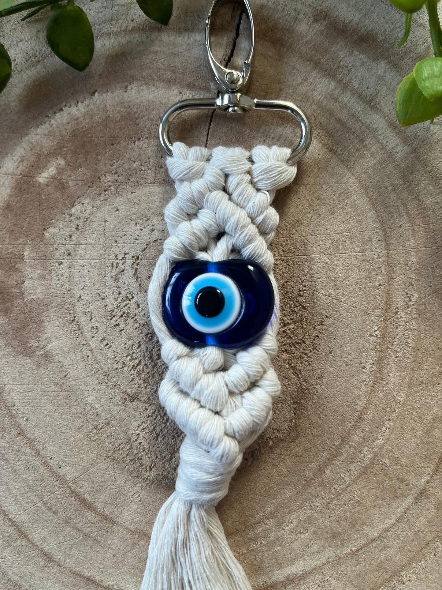 A Macra-Made-With-Love Evil eye keychain with a silver clasp is pictured. The handmade keychain is made of white braided cord and features a blue evil eye bead at its center, serving as a protection talisman. It is photographed on a wooden surface with some green leaves partially visible in the corners.