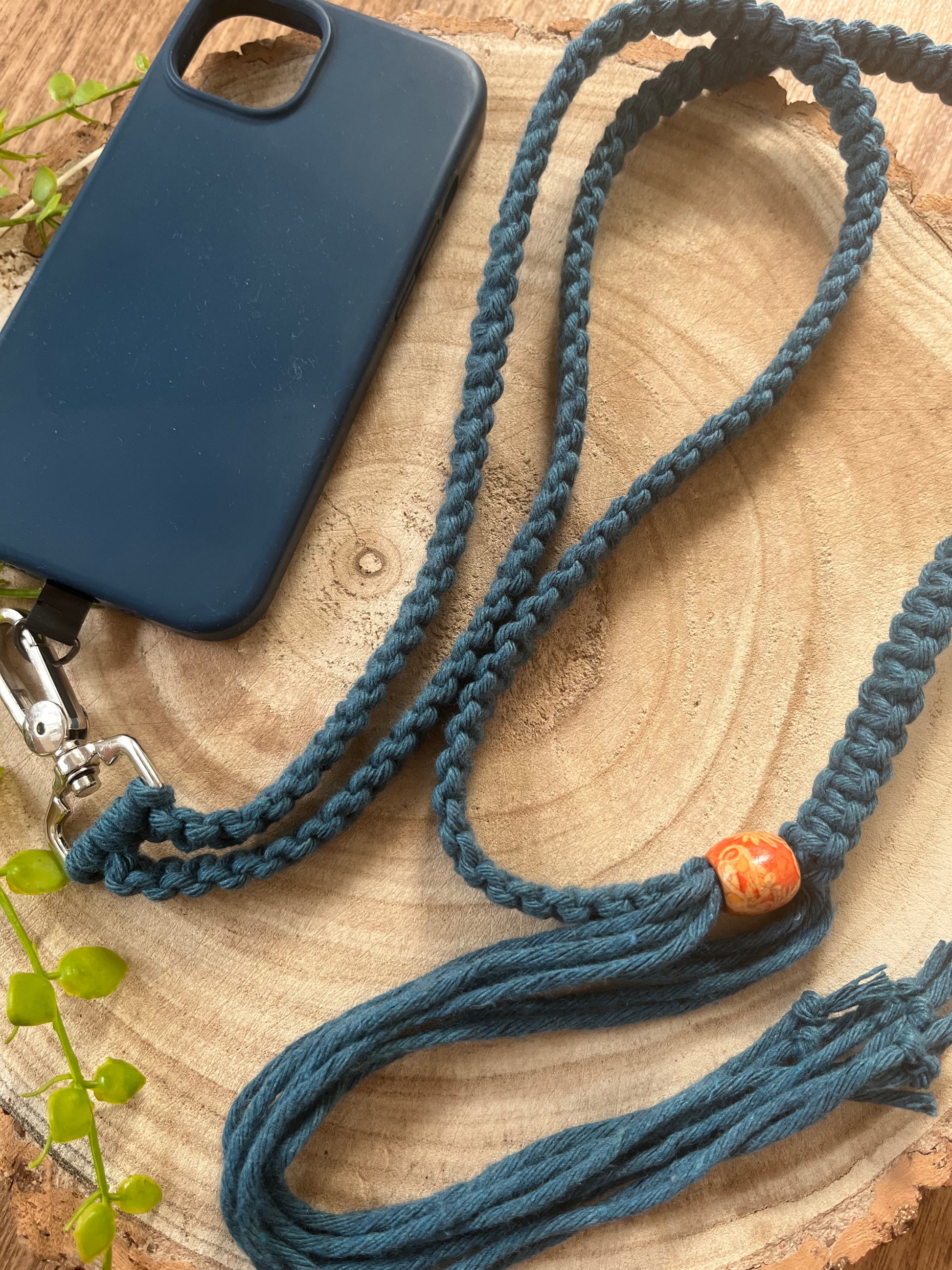 Lying on a wooden surface is a blue phone case adorned with Macra-Made-With-Love's Recycled cotton adjustable crossbody phone strap with universal tether. The rainbow strap, featuring a decorative orange bead and green vine leaves, adds an eco-friendly and stylish touch to the phone case.