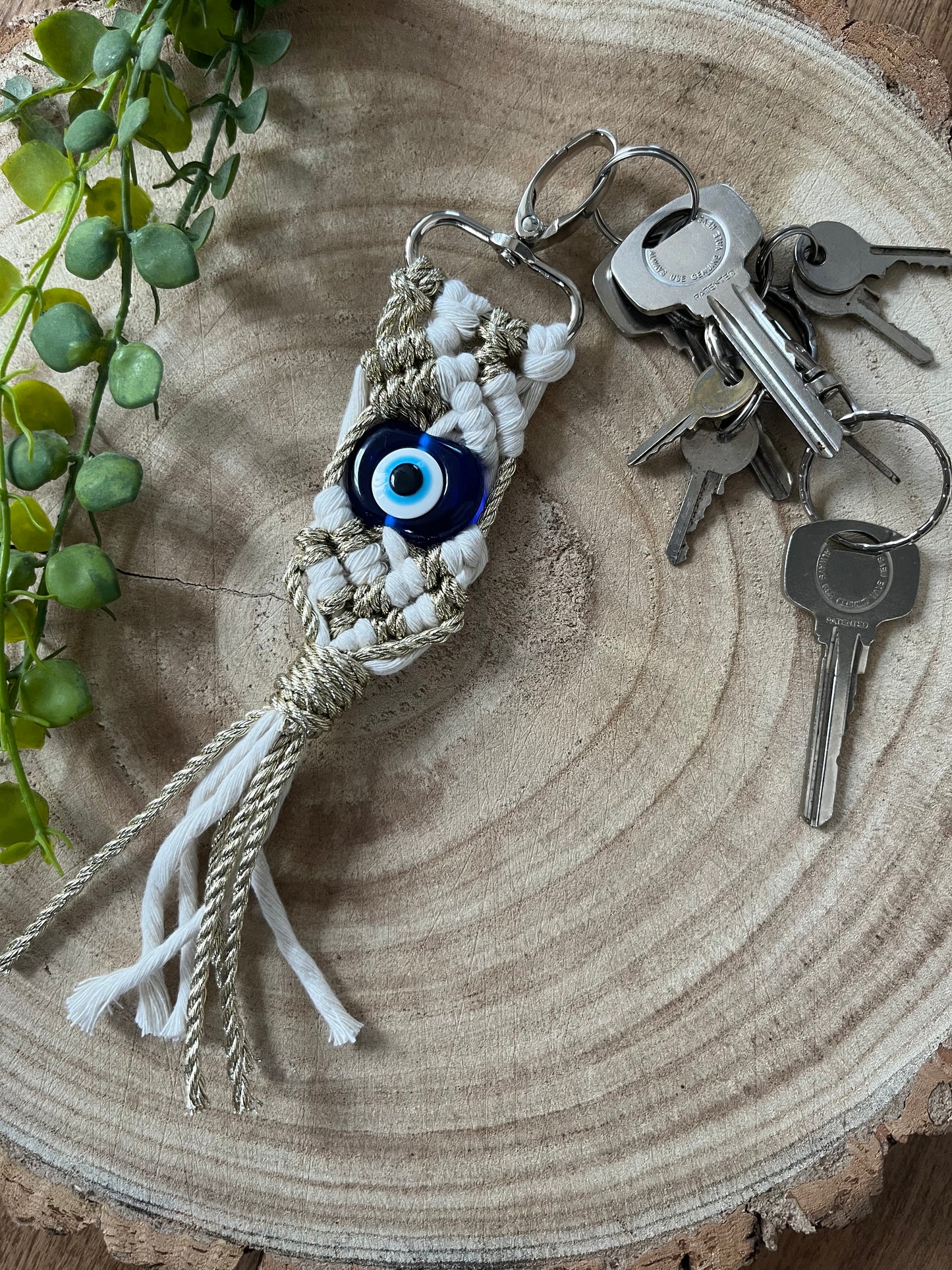 An Evil Eye keychain from Macra-Made-With-Love with an eye-shaped bead and macrame design lays on a wooden surface next to a set of keys. A green, leafy plant is partially visible on the side, adding a touch of nature to the composition.