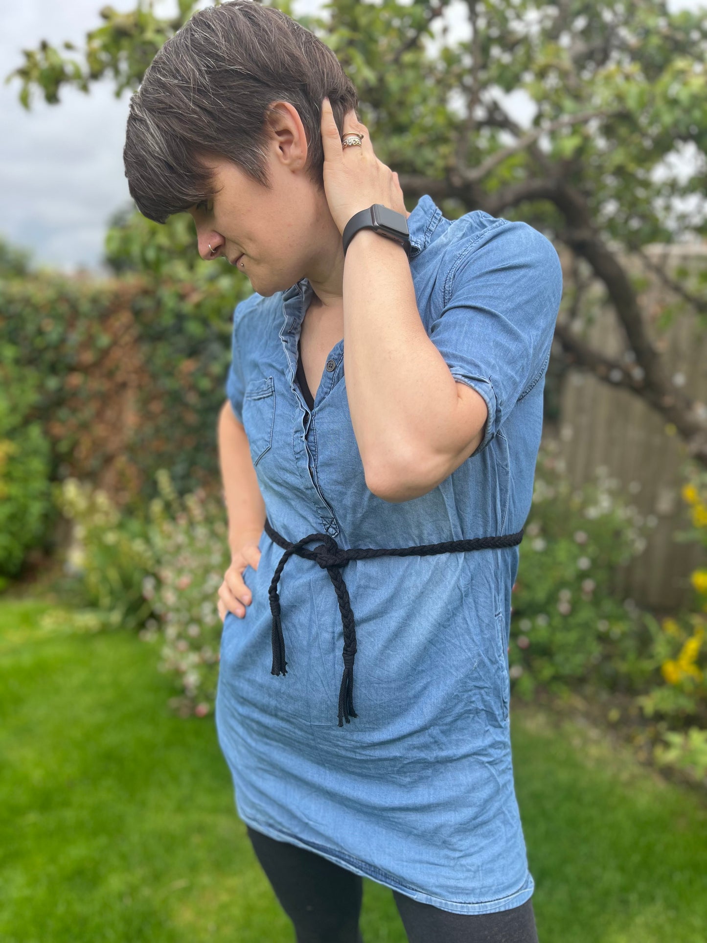 A person wearing a blue denim dress and a black wristwatch, along with an Eco-friendly Braided Macrame Belt from Macra-Made-With-Love, stands in a garden, one hand on the back of their neck and the other on their hip. The background features green grass, assorted flowers, and a tree.