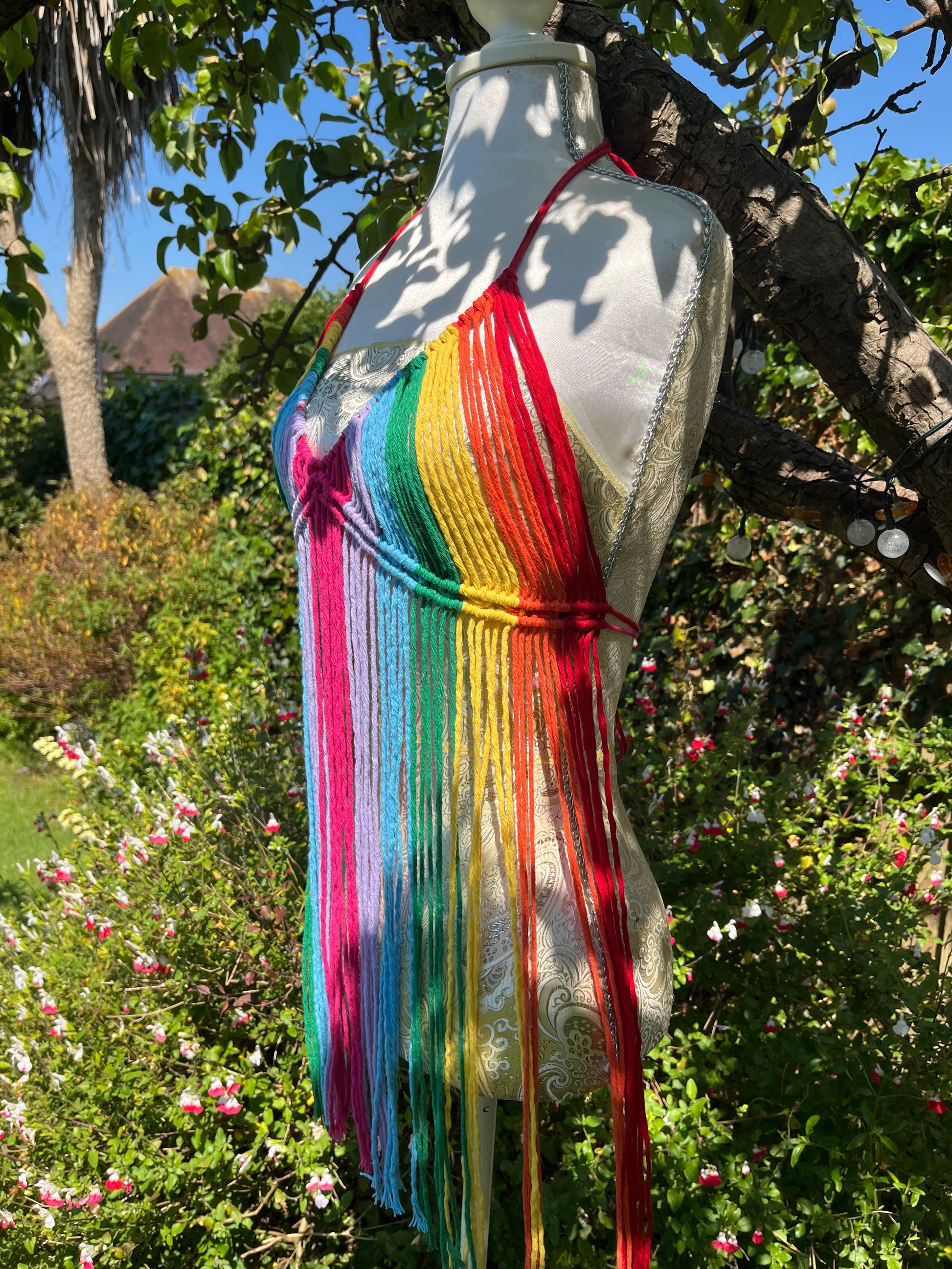 A mannequin wearing the Rainbow coverup top from Macra-Made-With-Love, a colorful, handmade macramé top with long fringe in rainbow colors is displayed outdoors in a lush garden, with a tree and vibrant greenery and flowers in the background. This bohemian dress makes the perfect festival outfit amidst nature's splendor.