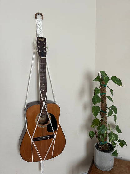 An acoustic guitar is hanging on the wall in a Macra-Made-With-Love Guitar Wall Mount made of decorative macramé. To the right of the guitar, there is a potted plant with green, heart-shaped leaves climbing a moss pole, placed on a wooden surface against a cream-colored wall.