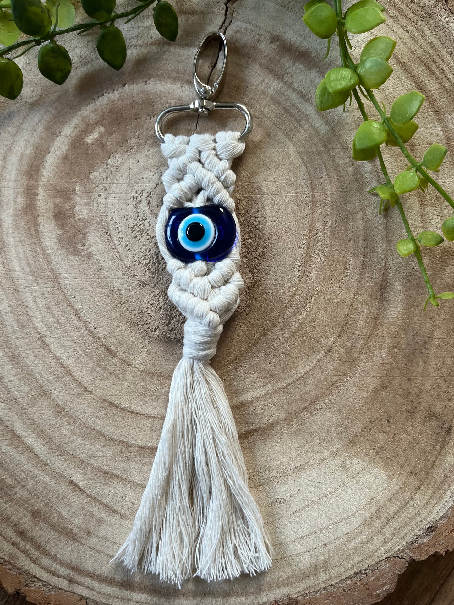 A handmade Evil eye keychain from Macra-Made-With-Love rests on a wooden surface, showcasing intricate knotted designs with a blue evil eye bead in the center and a tassel at the bottom. Green leaves are placed decoratively around this protection talisman.