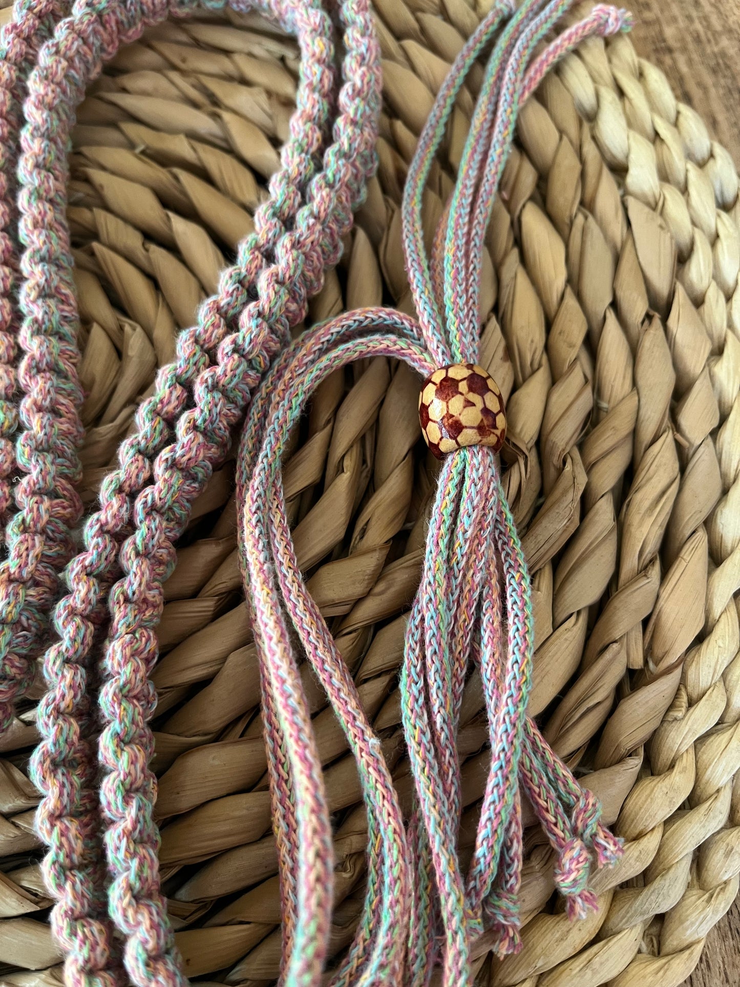 A close-up of the Recycled Cotton Adjustable Crossbody Phone Strap with Universal Tether from Macra-Made-With-Love, featuring a multicolored braided cord on a woven wicker surface. The end of the braid, crafted from eco-friendly recycled cotton, is secured with a patterned wooden bead. Loose strands extend from the bead, creating a decorative effect and highlighting this sustainable phone accessory.